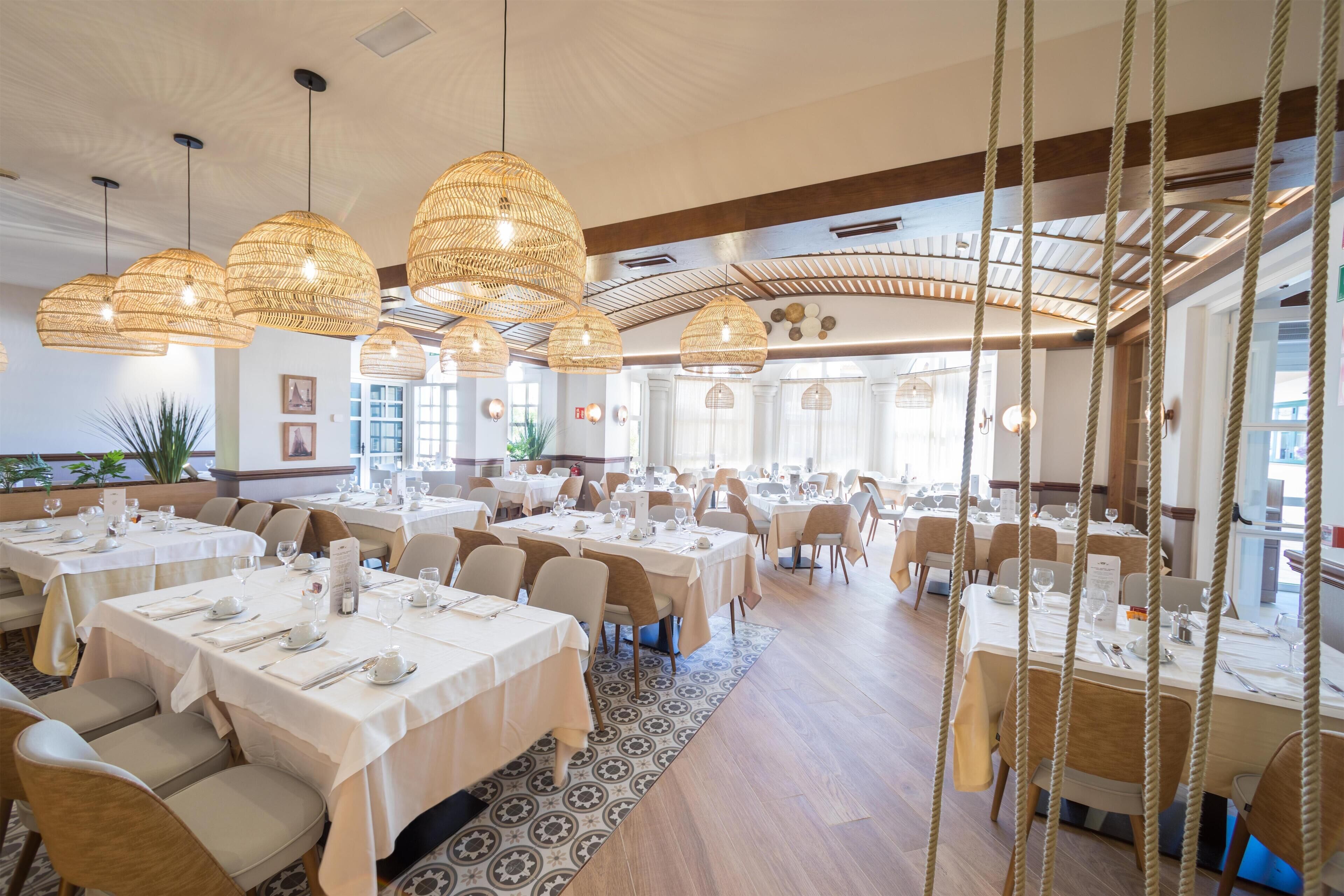 A spacious restaurant with multiple dining tables, wicker pendant lights, and a view of the outdoors.