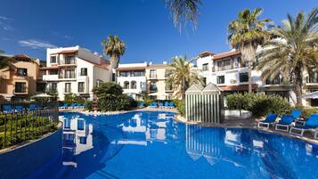 A swimming pool surrounded by palm trees and lounge chairs, with multi-story buildings in the background.