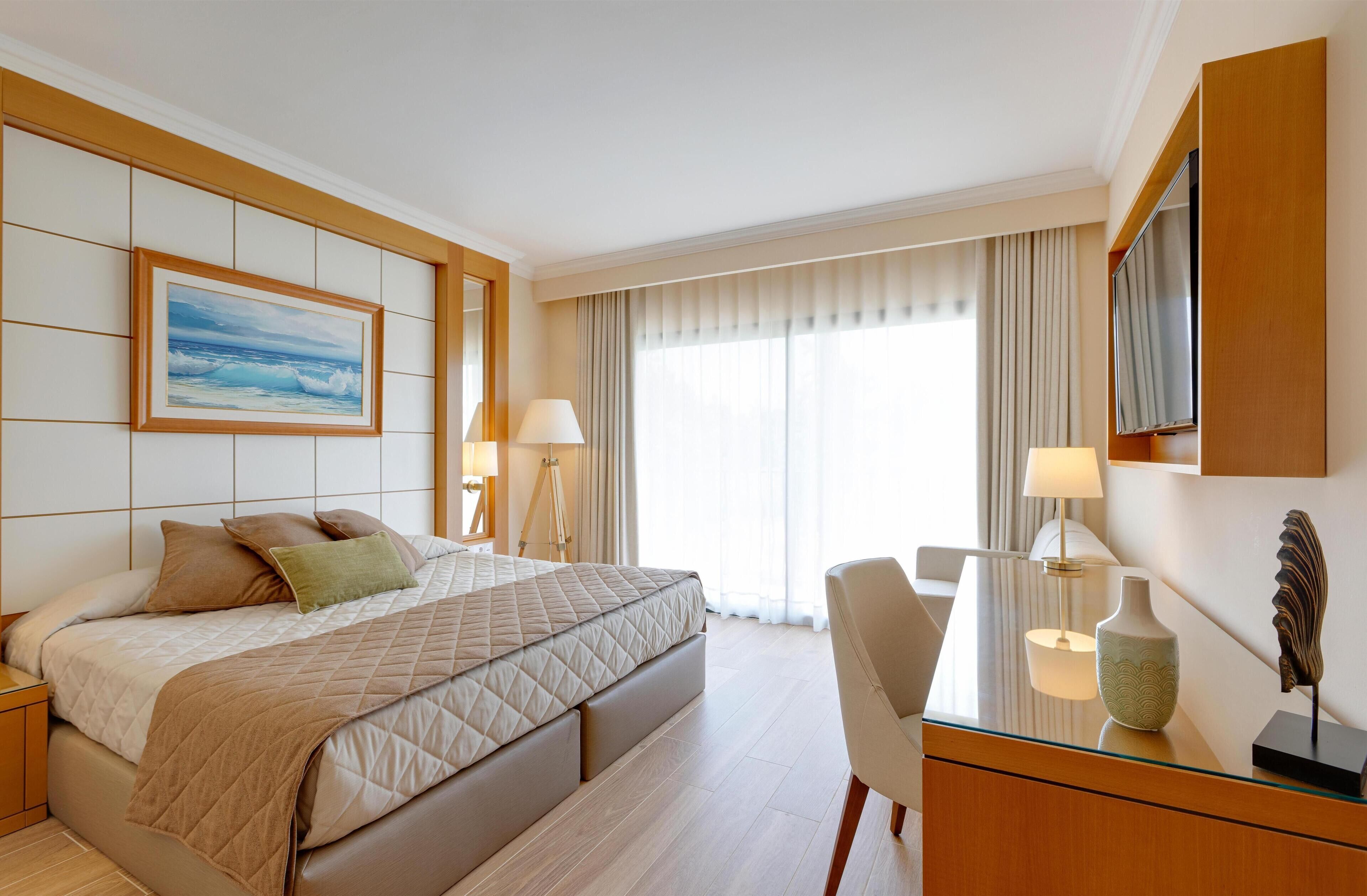 A hotel room with a large bed, a desk with a lamp, a television, and a painting on the wall.