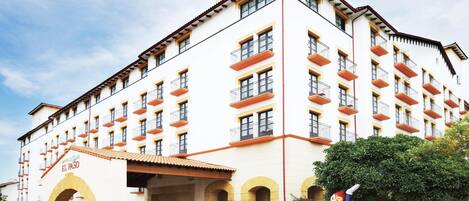 A multi-story hotel with a white facade, orange balconies, and a central entrance with an archway.