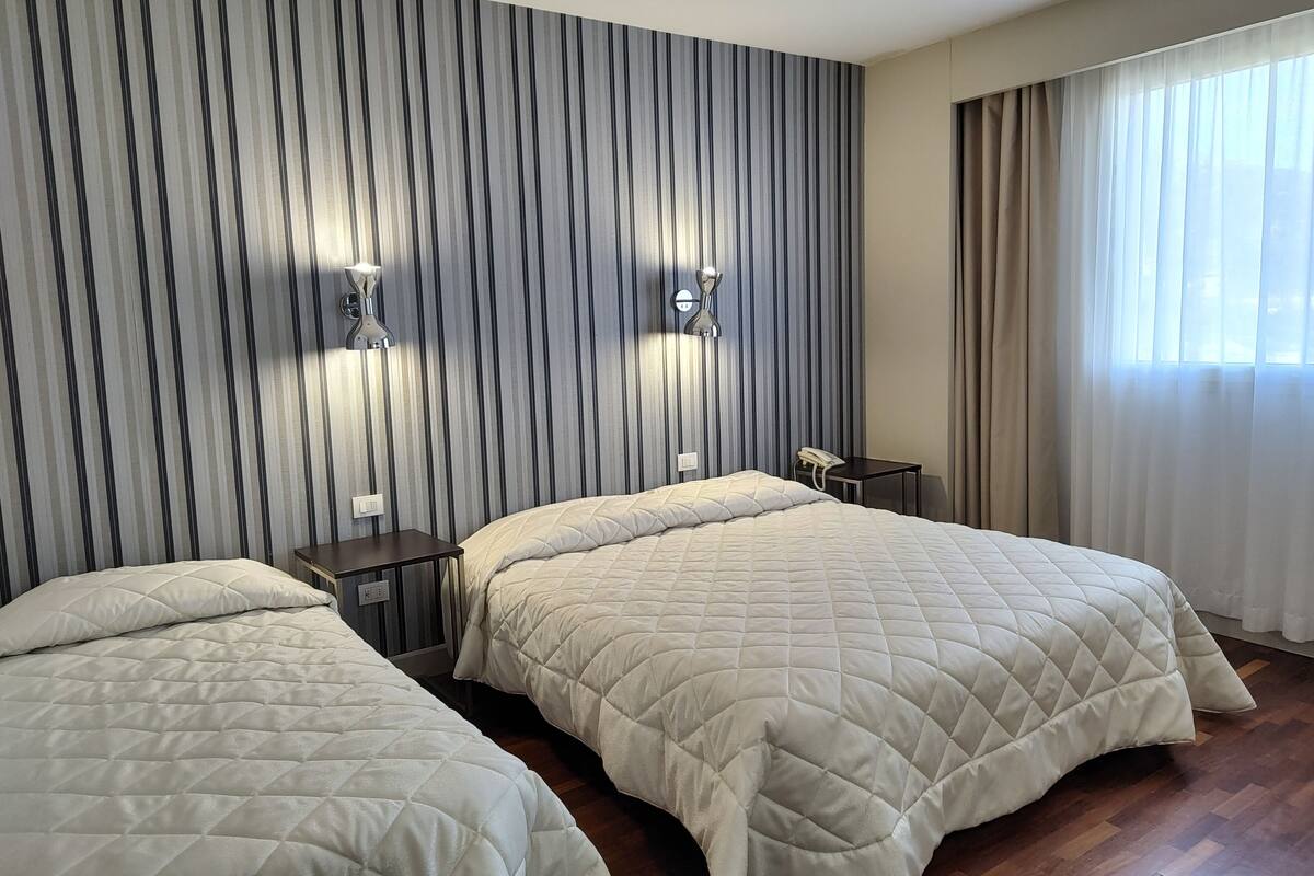 A hotel room with two beds, striped wallpaper, a nightstand with a lamp, and a window with curtains.