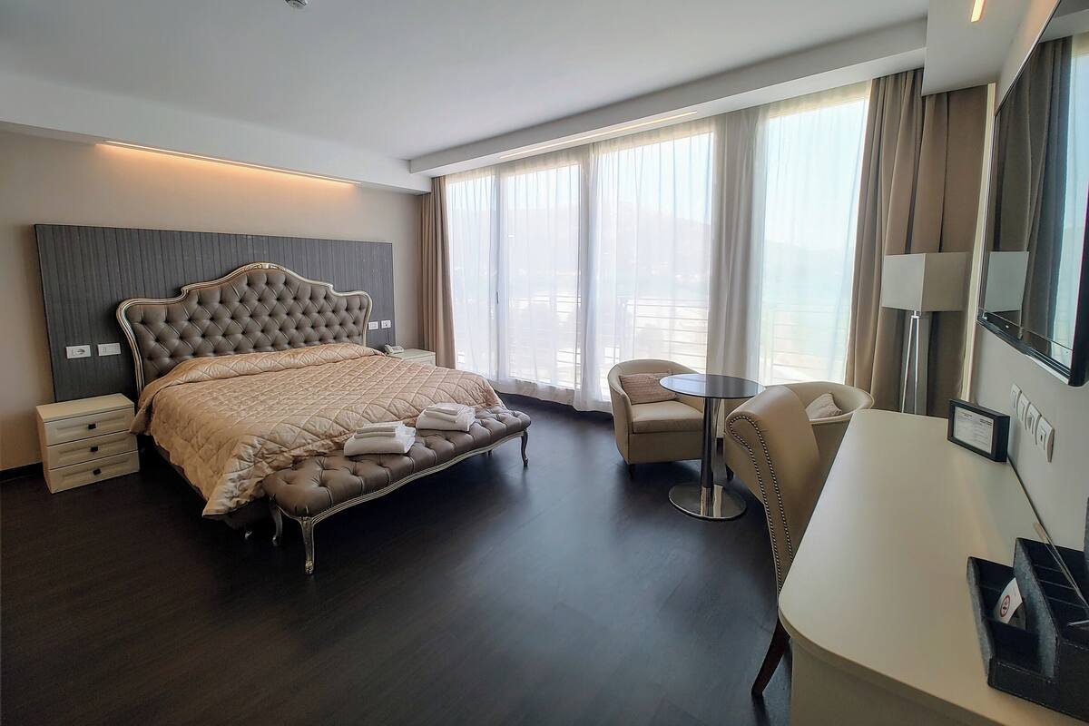 A modern hotel room with a large bed, a desk, a chair, and a balcony with curtains.