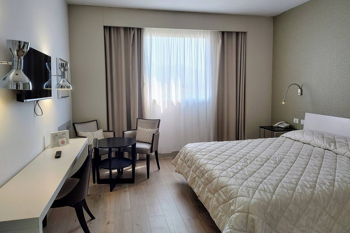A hotel room with a bed, a desk, two chairs, a TV, and a window with curtains.