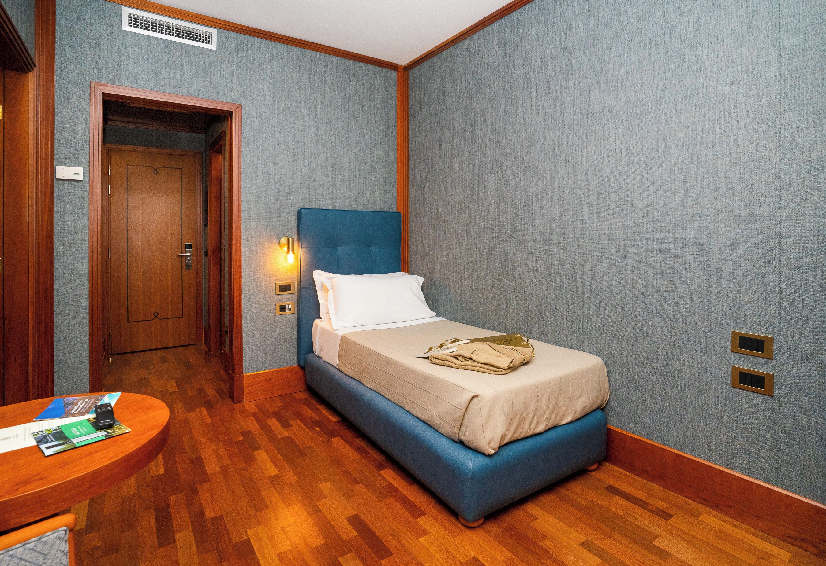 Una habitación de hotel con una cama azul, suelo de madera y una pequeña mesa con folletos.