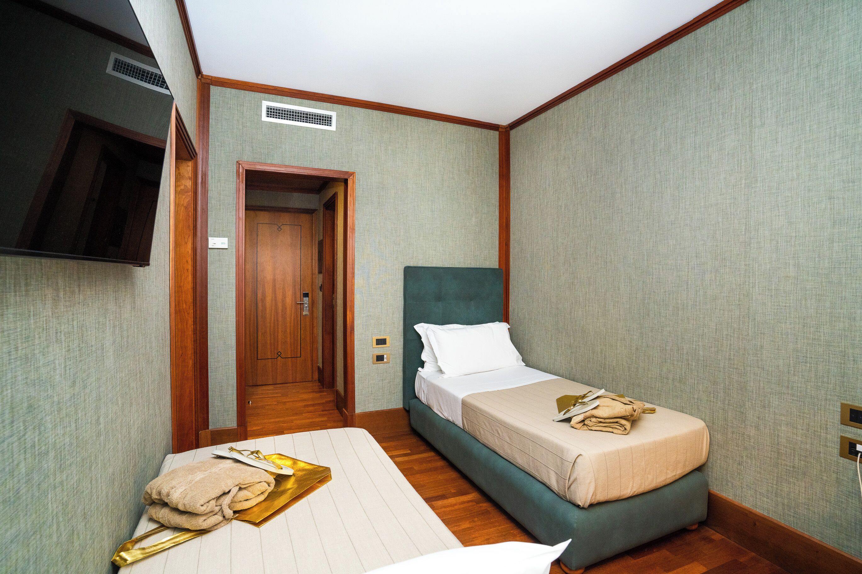 Habitación de hotel con una cama individual, una mesita de noche, un televisor montado en la pared y una puerta que da a otra habitación.
