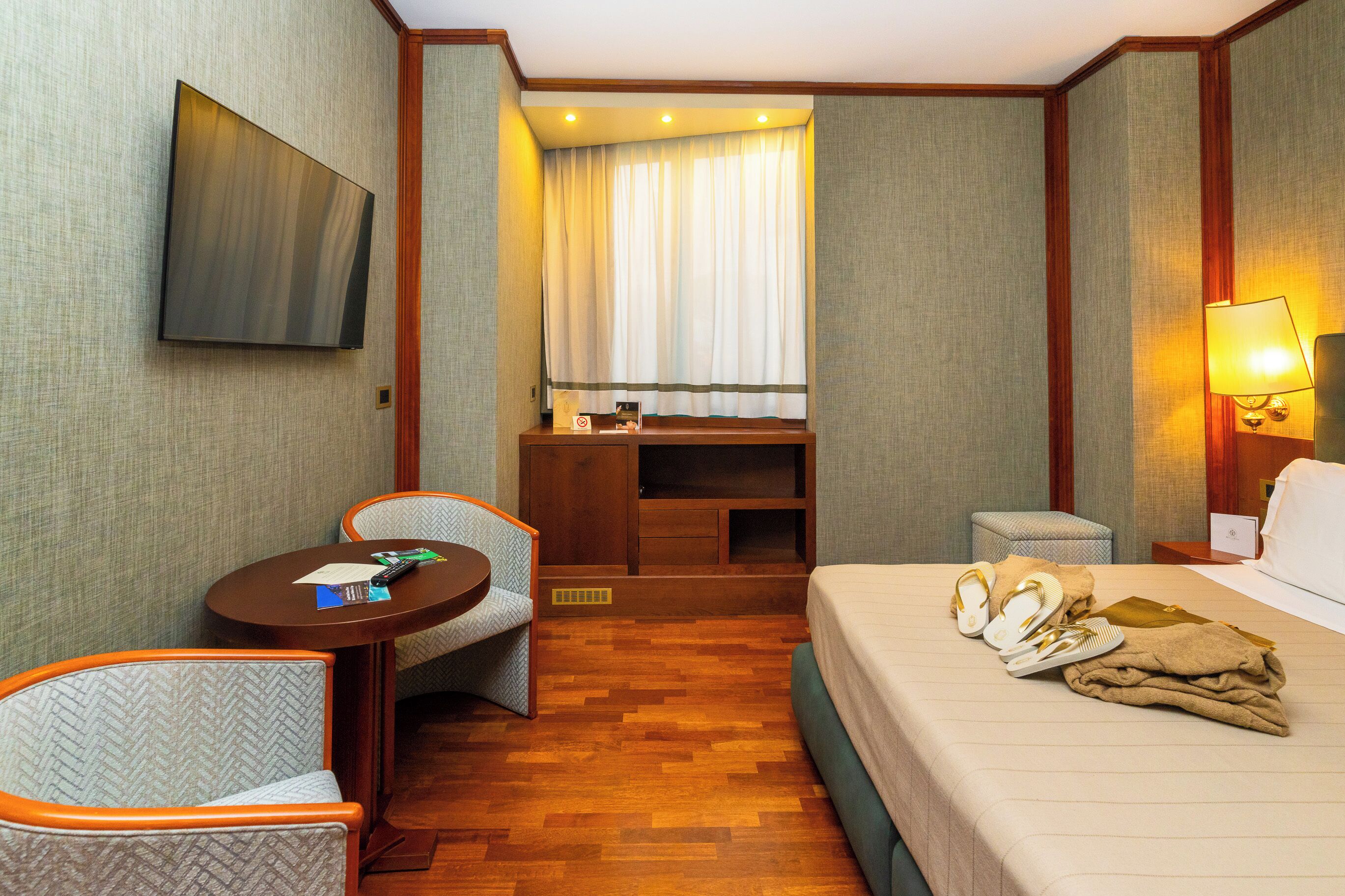 Habitación de hotel con cama, escritorio, silla, televisor y ventana con cortinas.