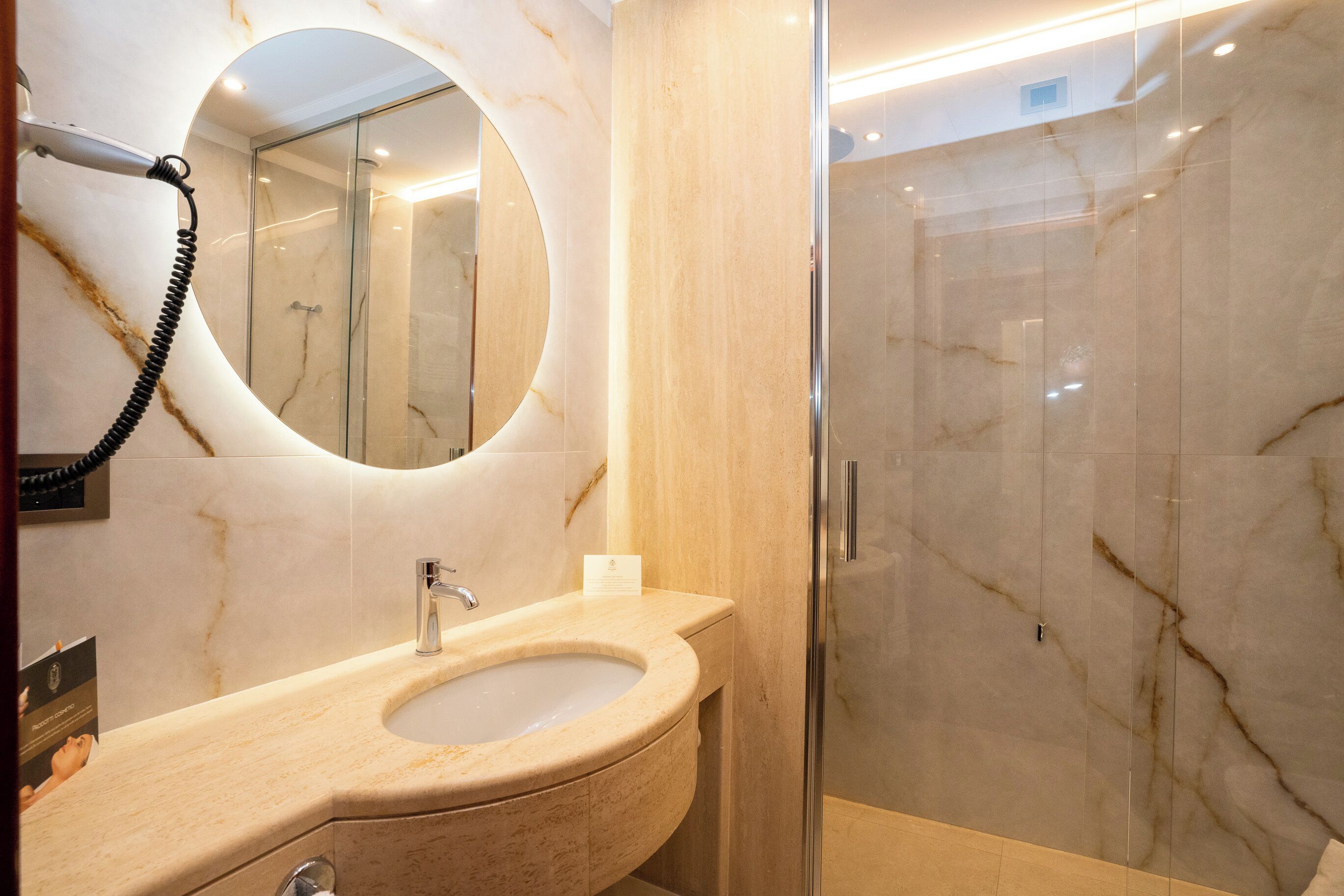 A modern bathroom with a marble sink, a round mirror, and a glass-enclosed shower.