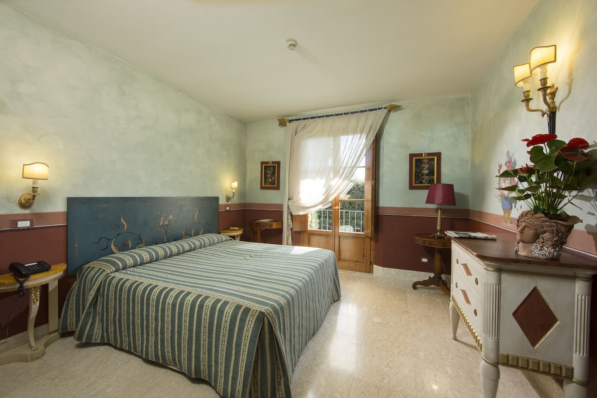 A hotel room with a large bed, a desk, a chair, a vanity with a mirror, and a window with curtains.