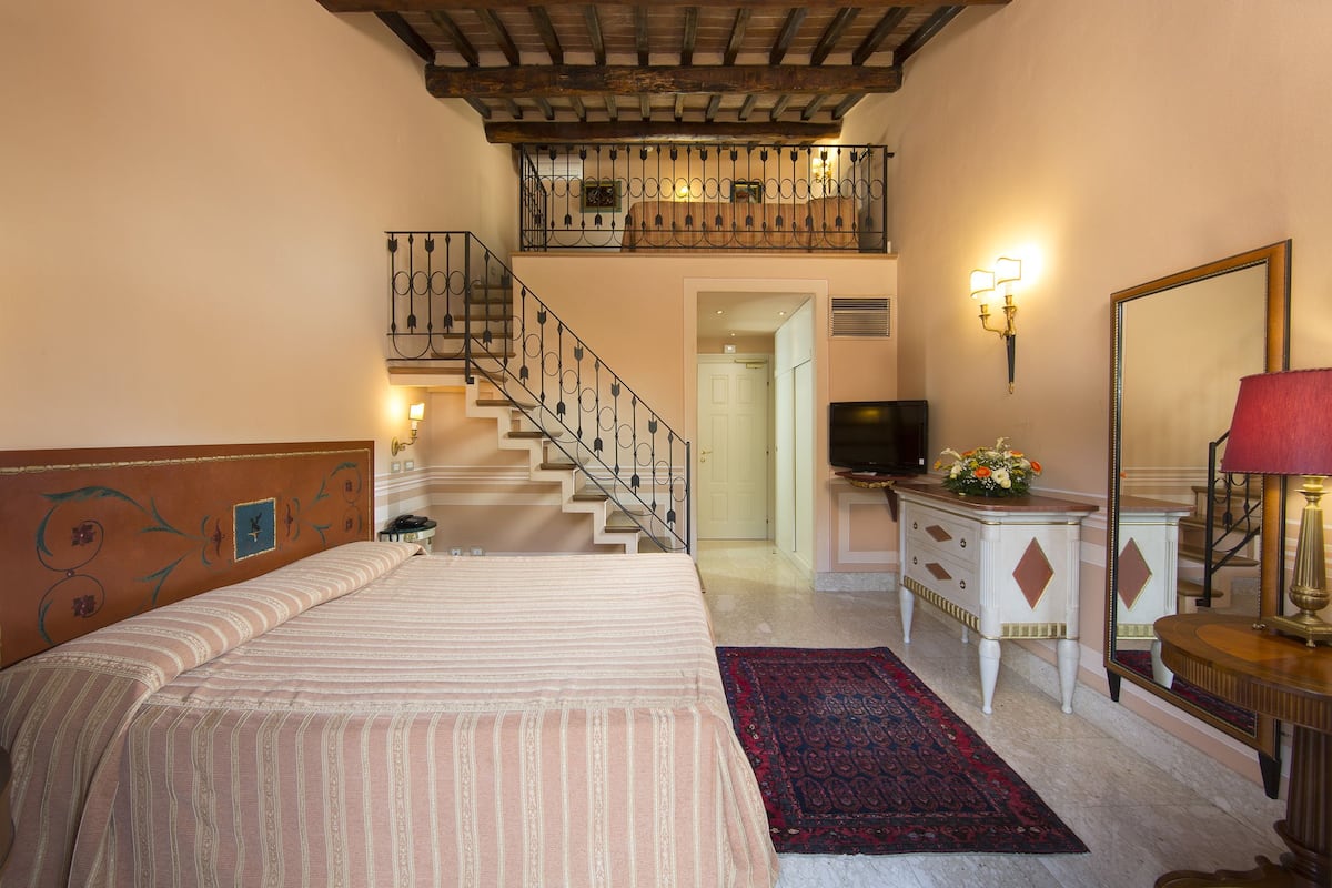 A bedroom with a bed, a desk, a dresser, a mirror, a staircase, and a balcony.