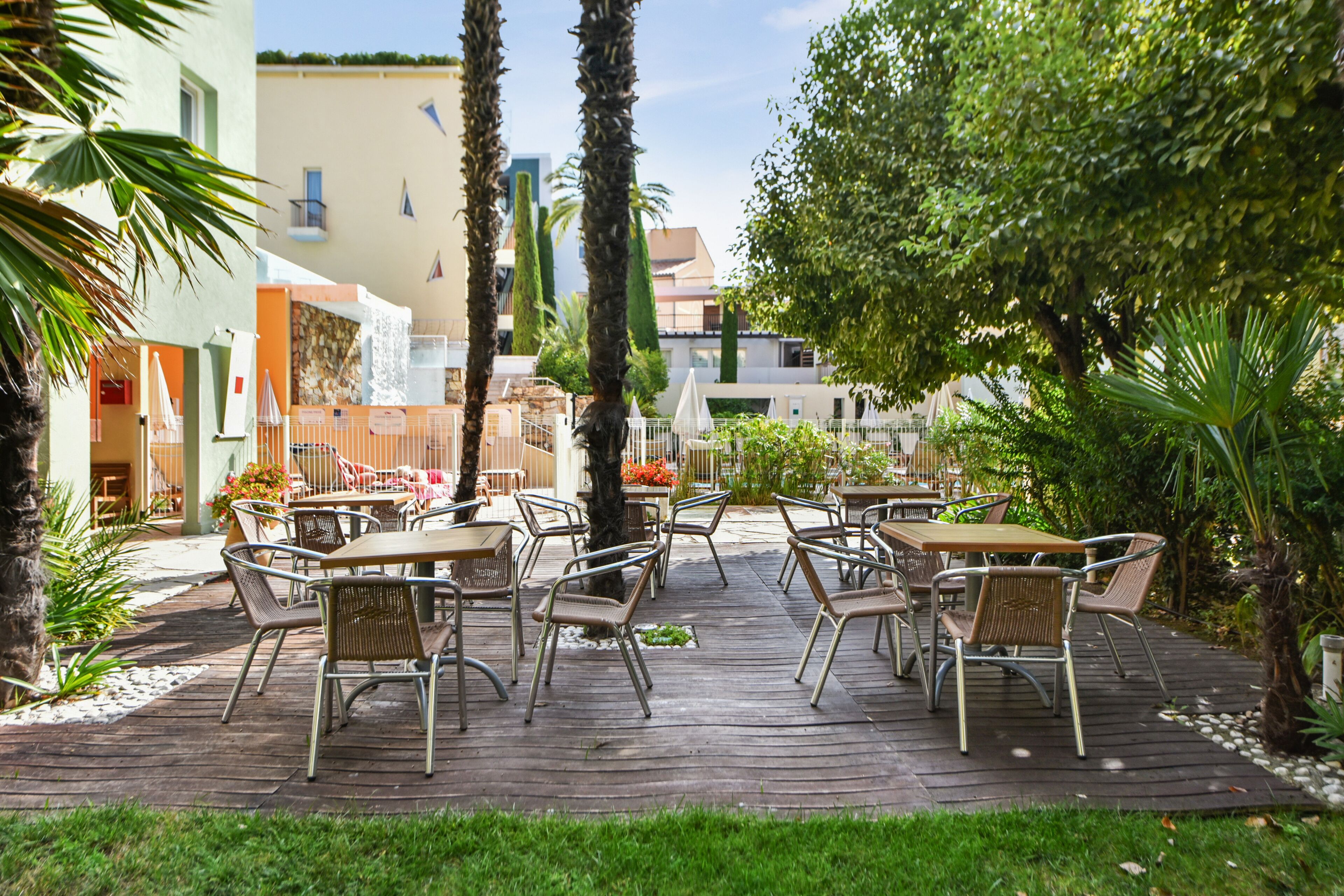 Un patio al aire libre con mesas y sillas de madera, rodeado de vegetación y edificios.