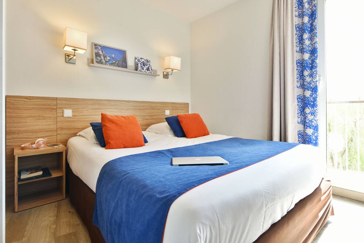 A hotel room with a wooden headboard, a bed with blue and white bedding, two orange pillows, a bedside table with a lamp, and a window with a blue curtain.