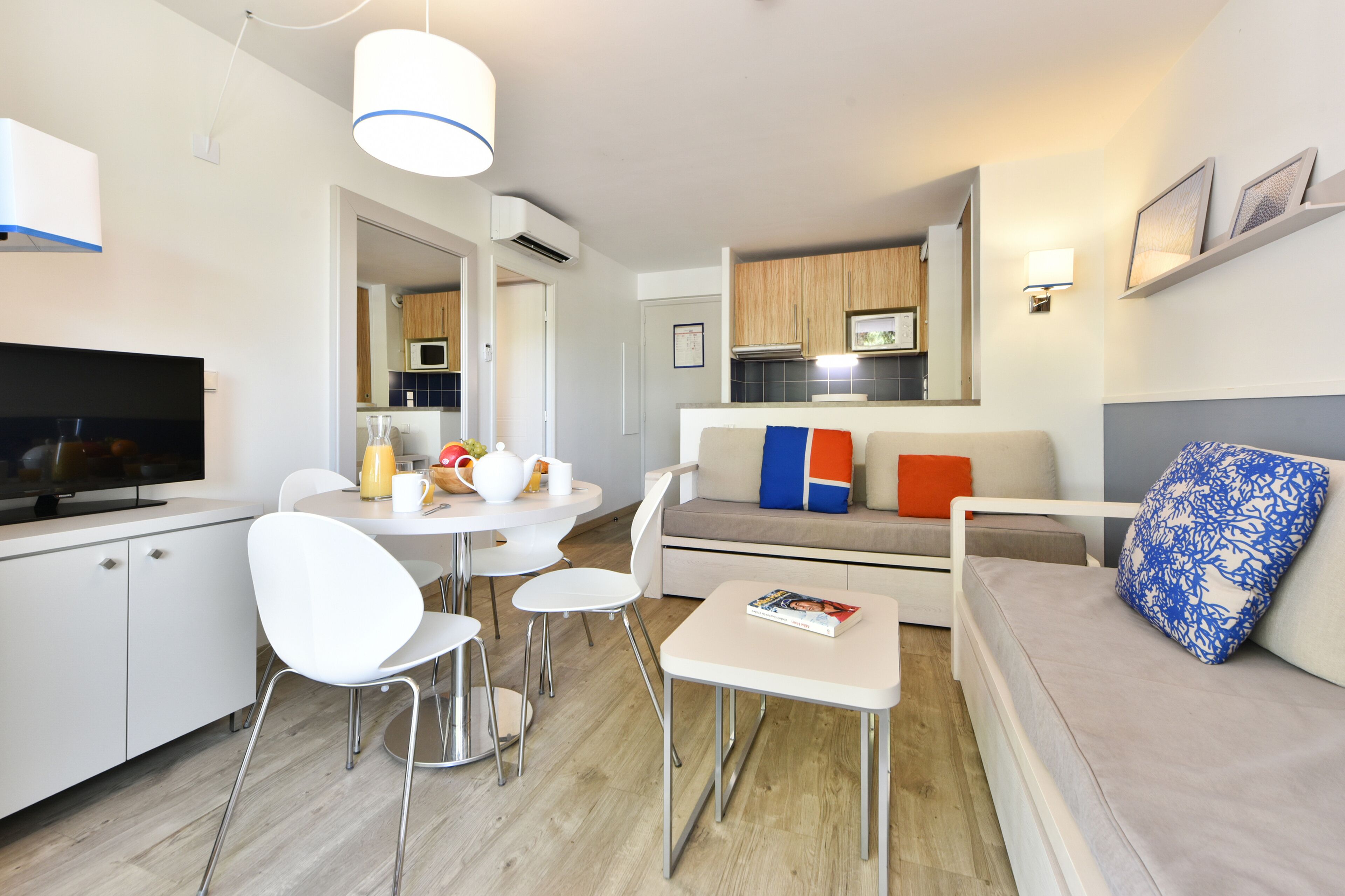 Una sala de estar moderna con un televisor de pantalla plana, una mesa de comedor con sillas, un sofá con cojines coloridos, y una zona de cocina al fondo.