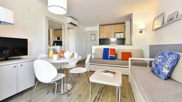 Una sala de estar moderna con un televisor de pantalla plana, una mesa de comedor con sillas, un sofá con cojines coloridos, y una zona de cocina al fondo.