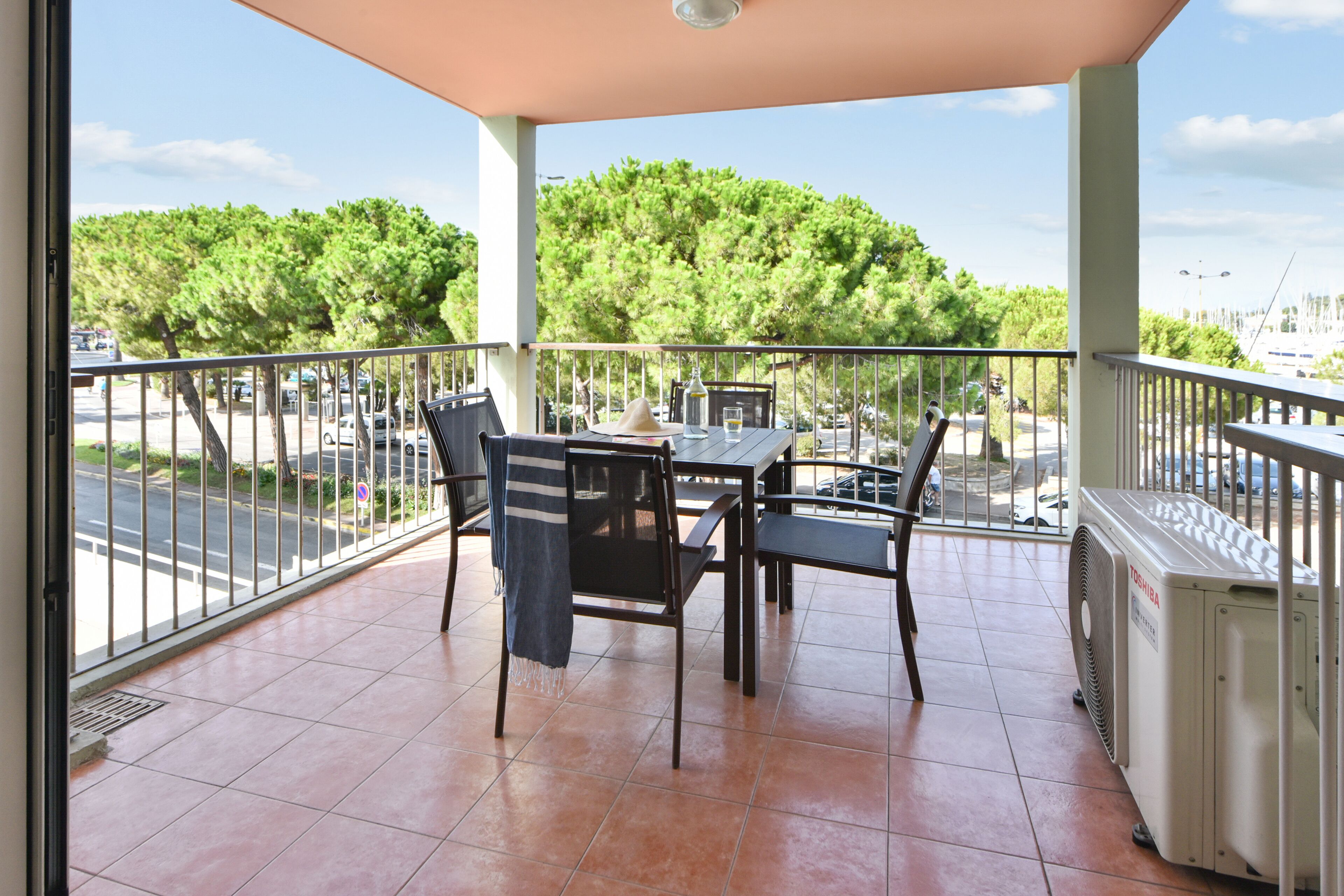 A balcony with a table and chairs, a fan, and a view of trees and a road.