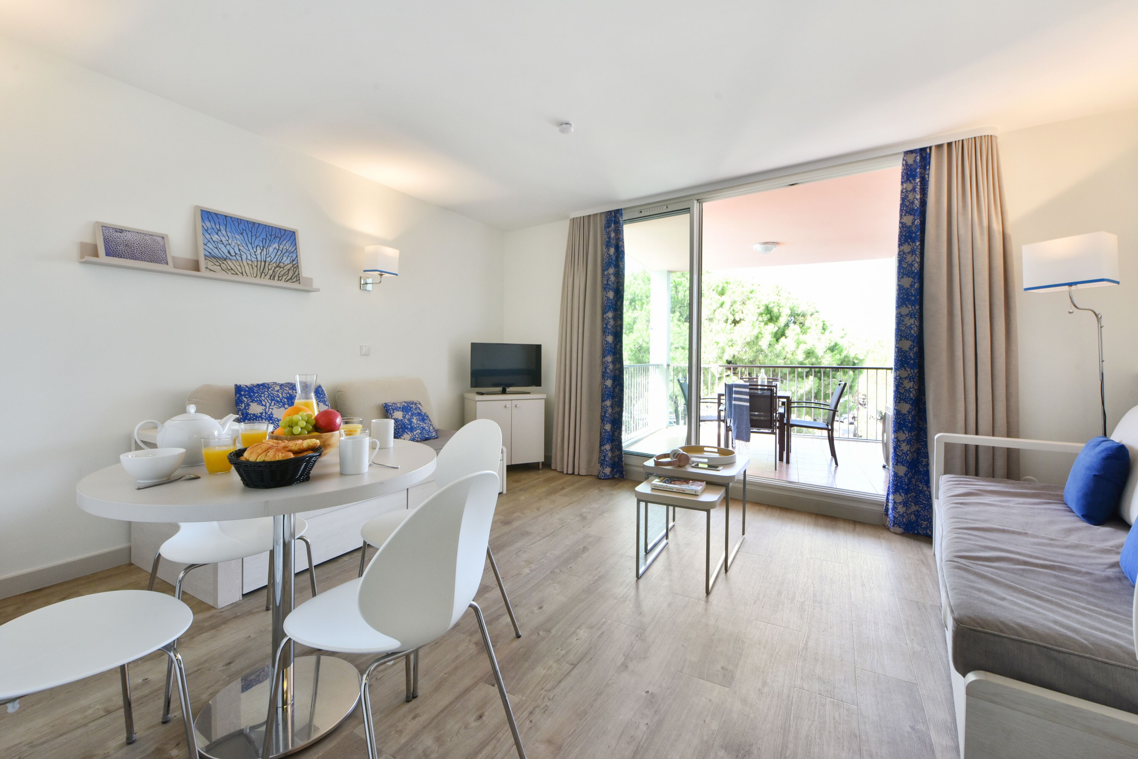 A modern hotel room with a dining area, a sofa, a TV, and a balcony with a view of greenery.