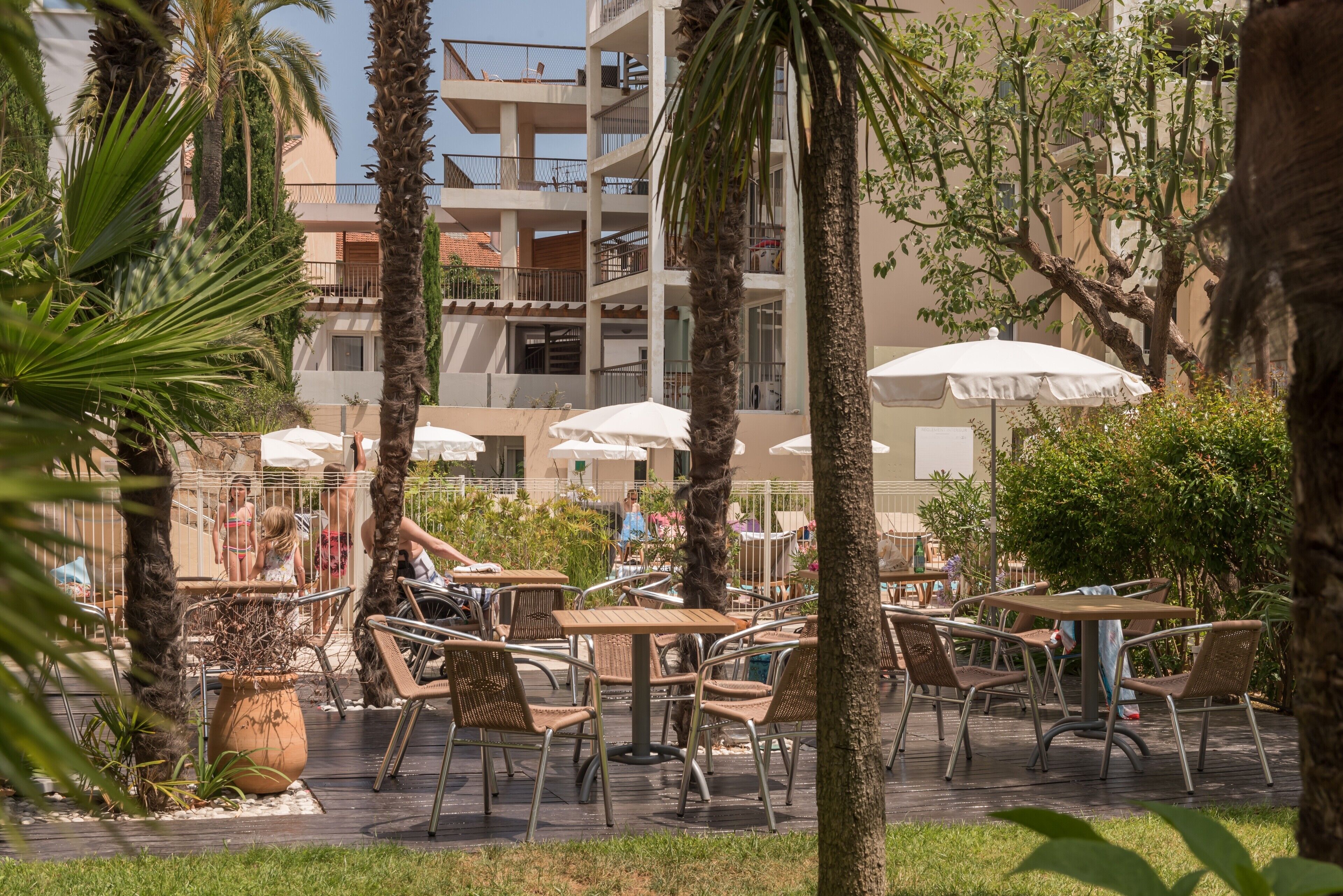 Outdoor seating with umbrellas, palm trees, and a building with balconies in the background.