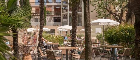 Outdoor seating with umbrellas, palm trees, and a building with balconies in the background.