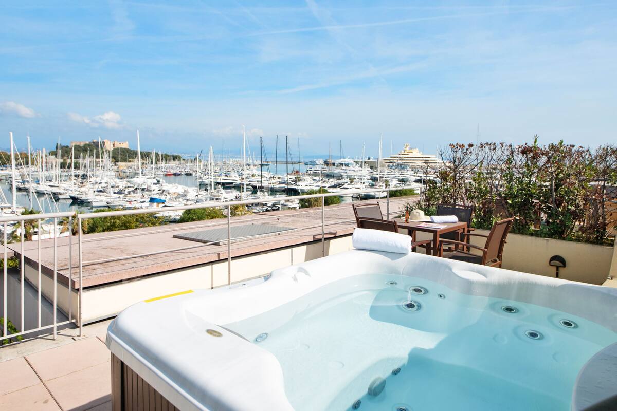 A rooftop terrace with a hot tub, overlooking a marina with numerous boats and a castle in the distance.