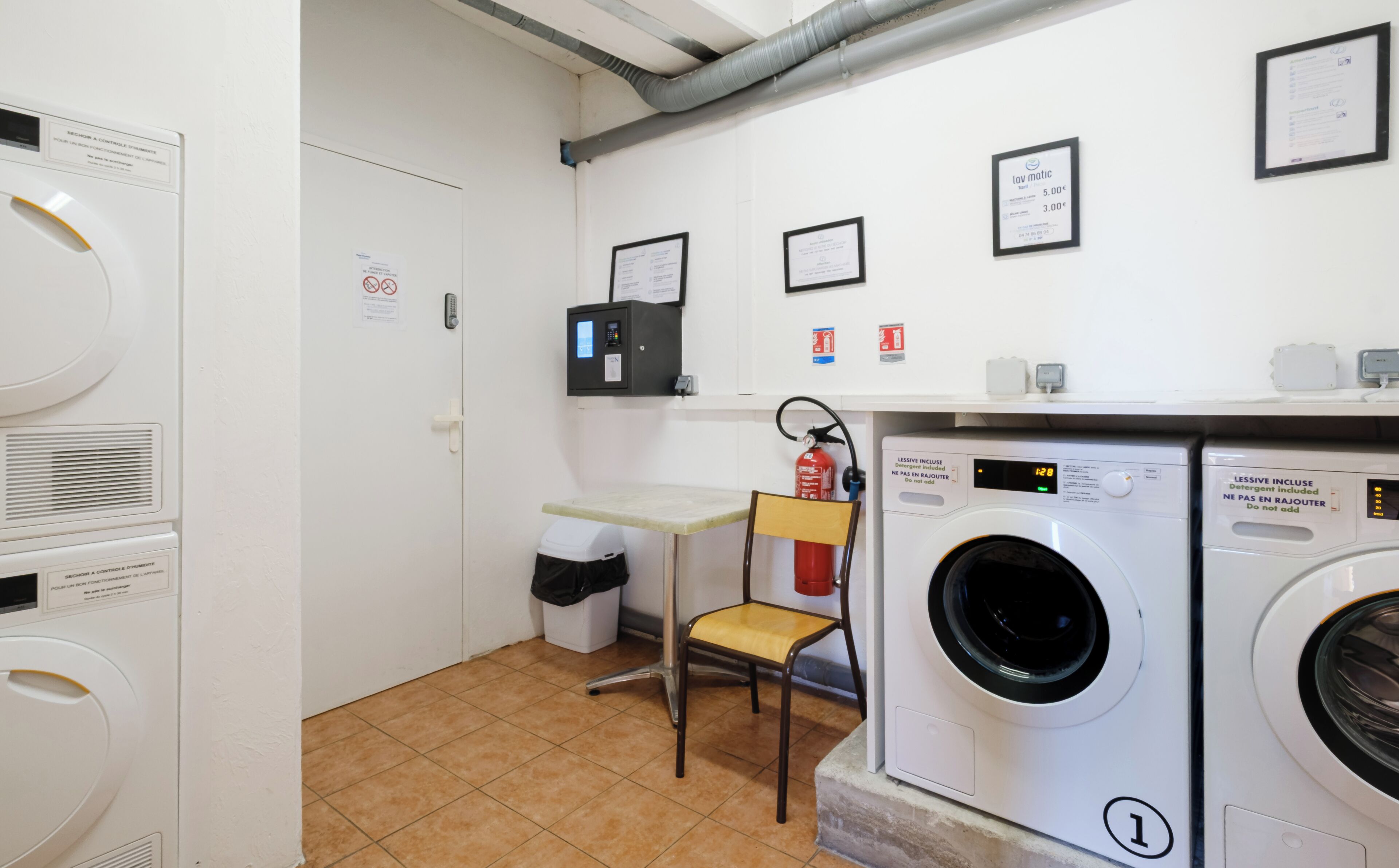 Een wasserette met een wasmachine, een droger, een stoel, een brandblusser en een bedieningspaneel.