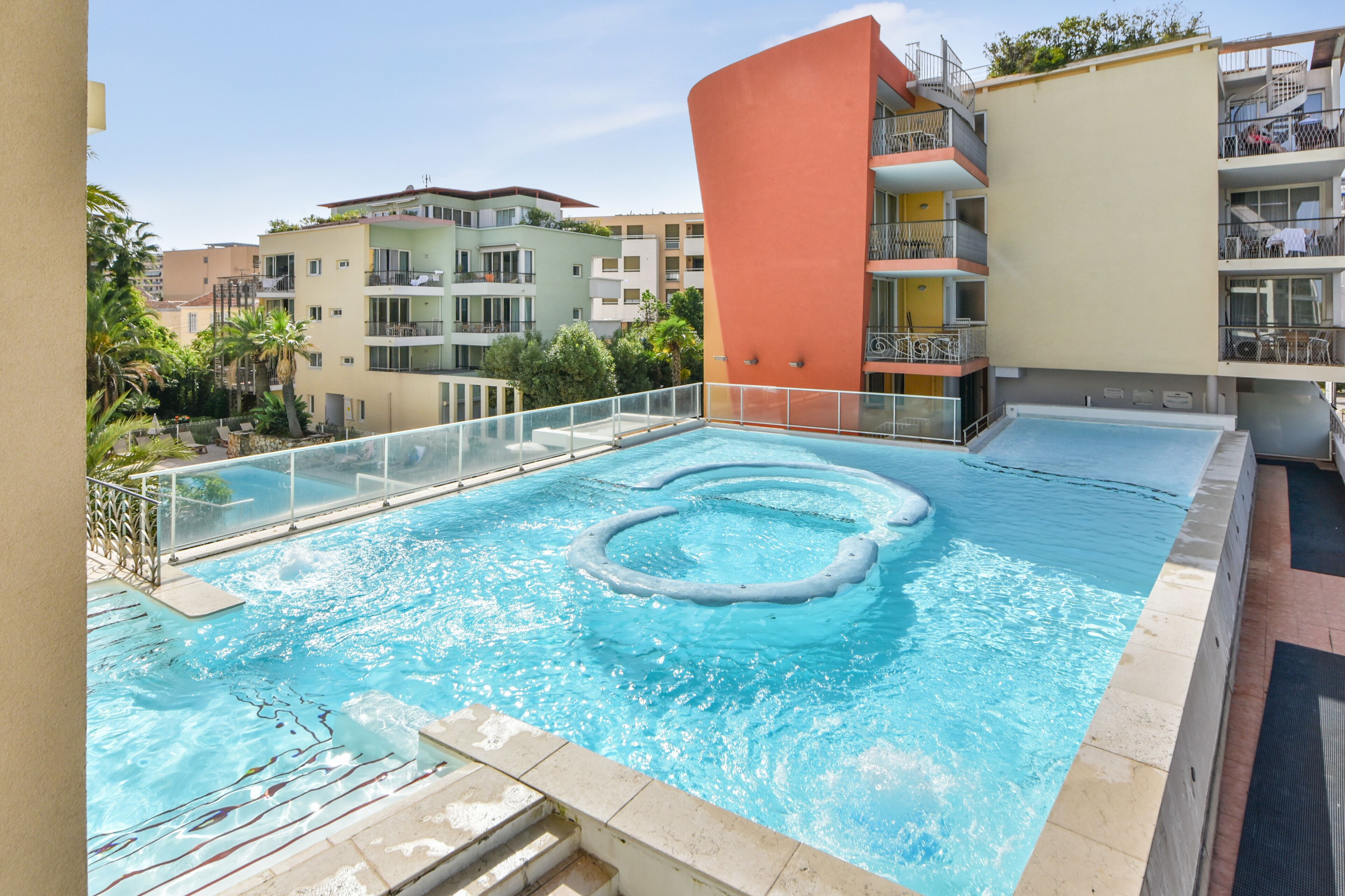 A rooftop pool with a circular hot tub, surrounded by a glass railing, set against a backdrop of multi-story buildings.