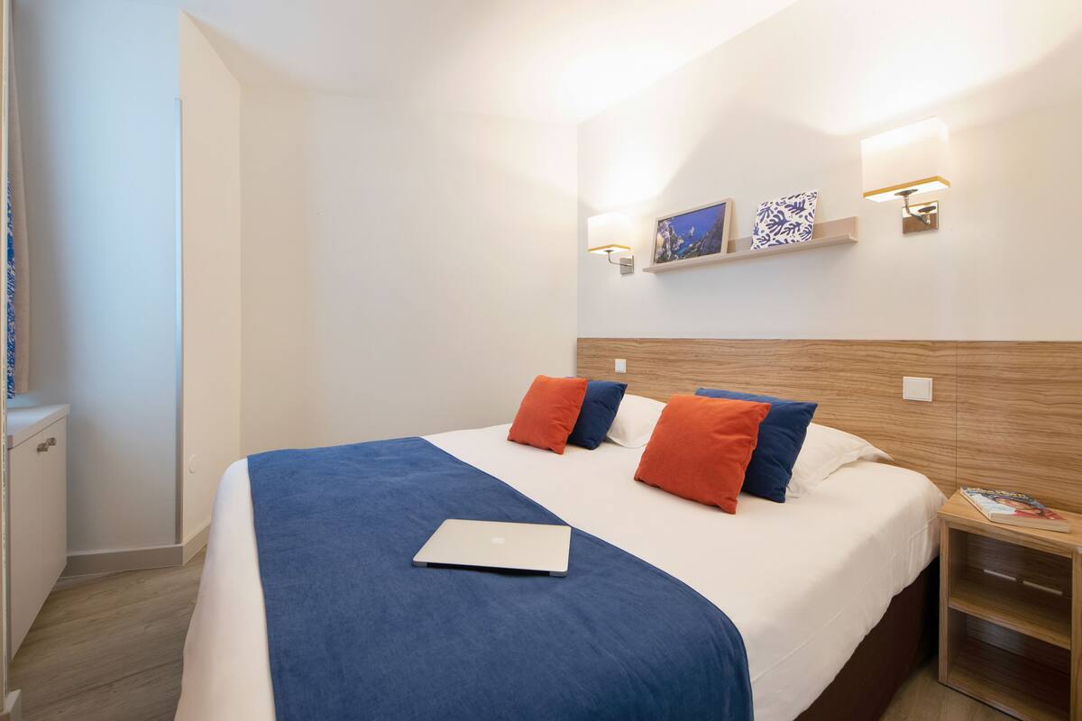 A neatly made bed with a laptop, a wooden headboard, and two wall-mounted light fixtures.
