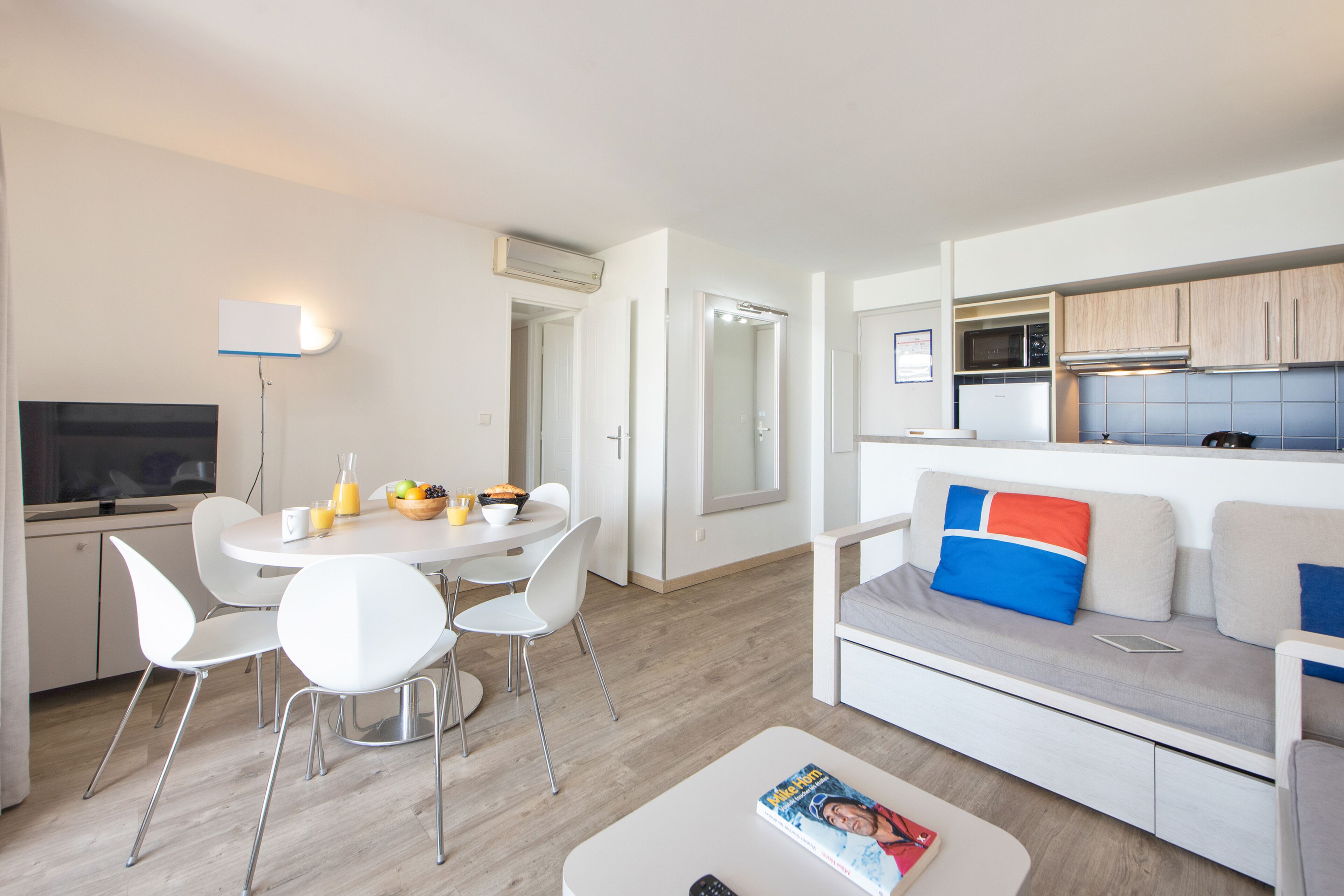 Una sala de estar moderna con un sofá blanco, una mesa de comedor con sillas, una zona de cocina con armarios y electrodomésticos, y un televisor.