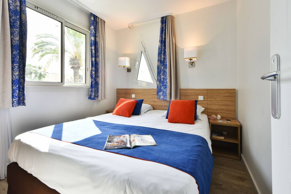 A hotel room with a large bed, two nightstands, a window with curtains, and a wall-mounted lamp.