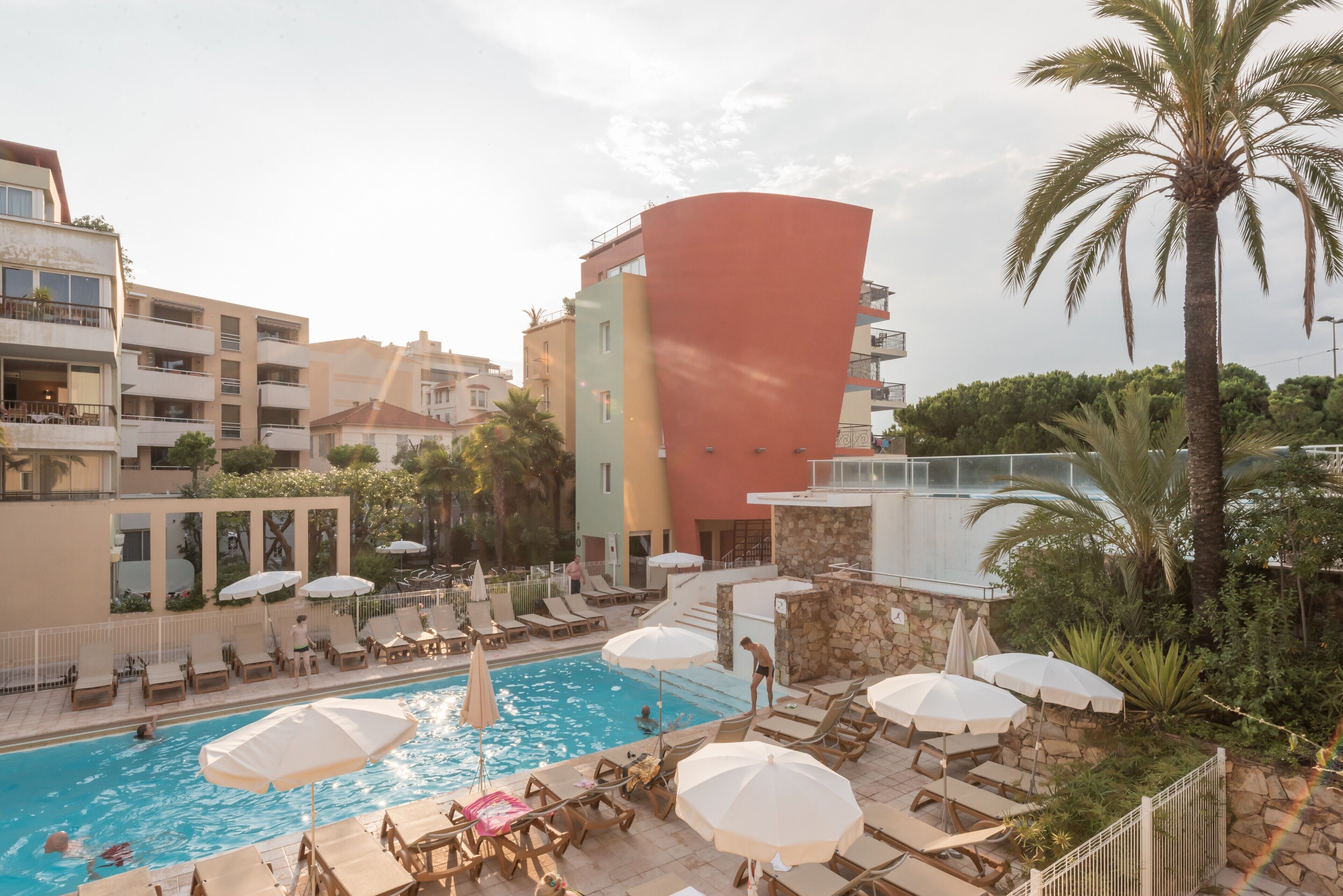 Un área de piscina con sillas de descanso y sombrillas, rodeada de edificios y palmeras.