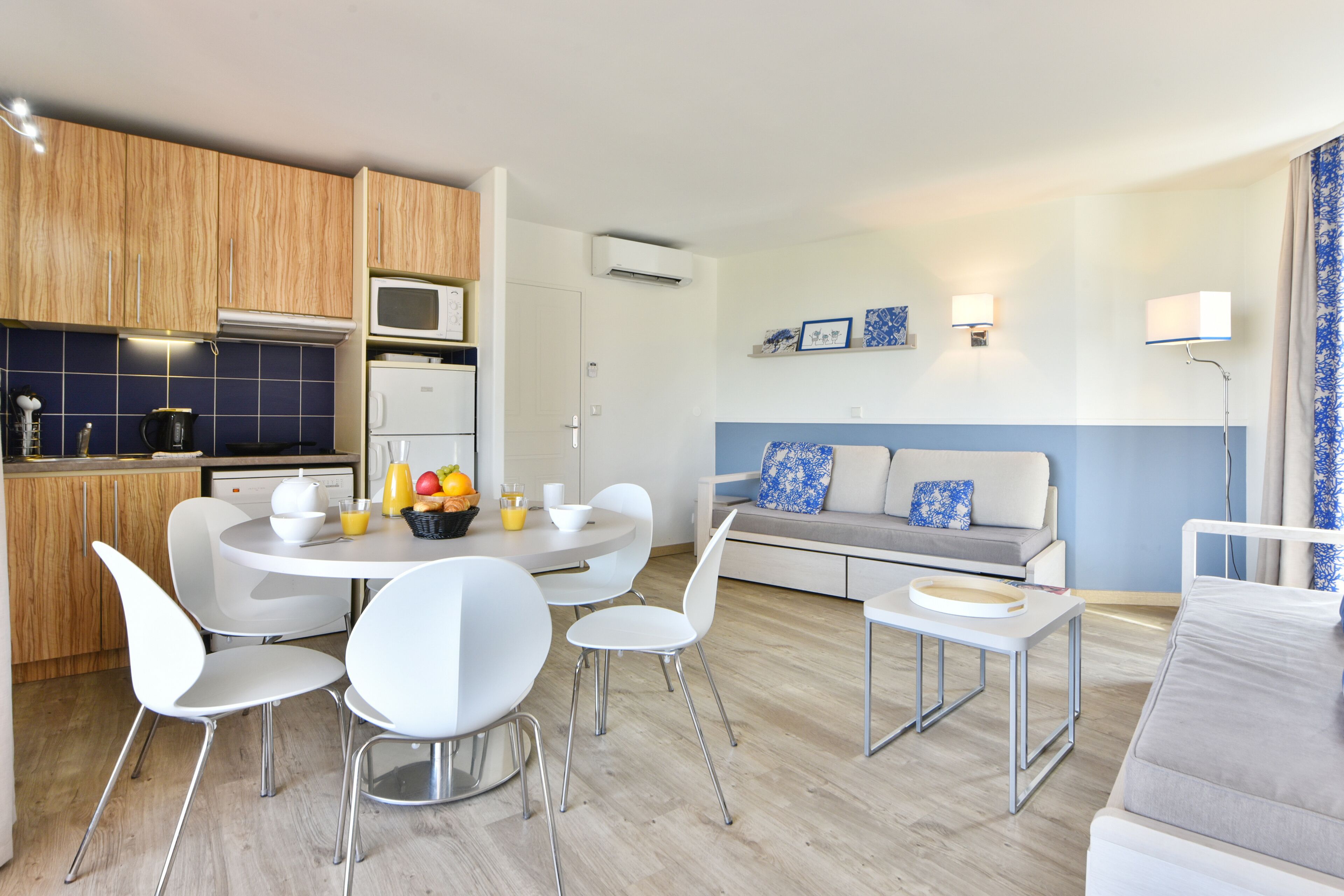 Une cuisine moderne avec des appareils électroménagers blancs, un coin repas avec des chaises blanches et un salon avec un canapé gris et une décoration bleue.