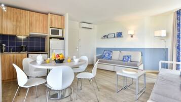 Une cuisine moderne avec des appareils électroménagers blancs, un coin repas avec des chaises blanches et un salon avec un canapé gris et une décoration bleue.