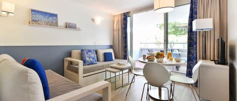 A modern hotel room with a sofa, a dining table with chairs, a small kitchen area, and a balcony with curtains.