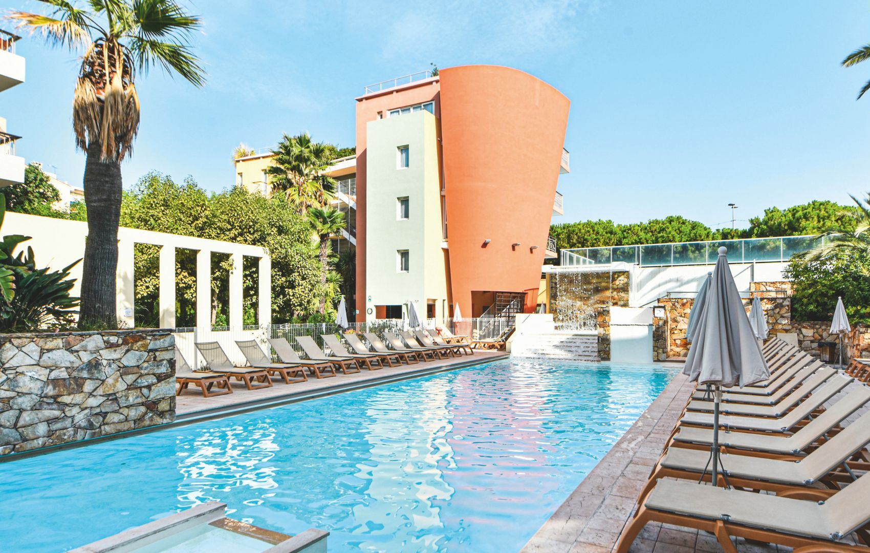 A swimming pool with lounge chairs and umbrellas, surrounded by palm trees and a building with a curved facade.