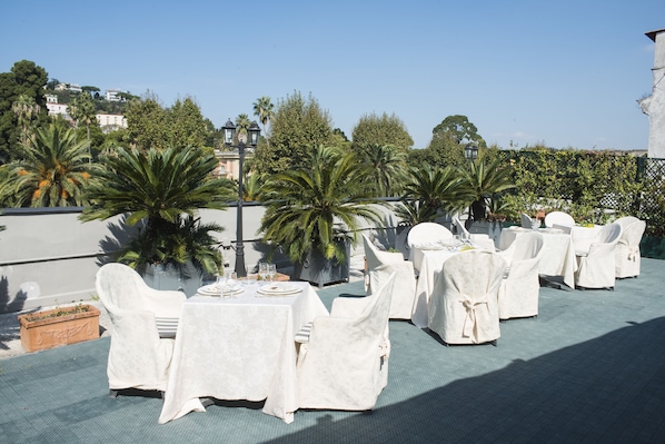 Un área de comedor al aire libre con mesas y sillas, rodeada de follaje y un cielo despejado.