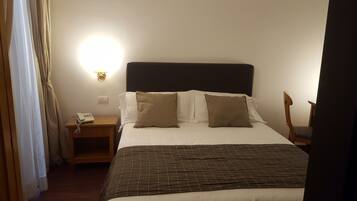 Una camera d'albergo con un letto, un comodino con il telefono, una lampada a parete e una sedia.