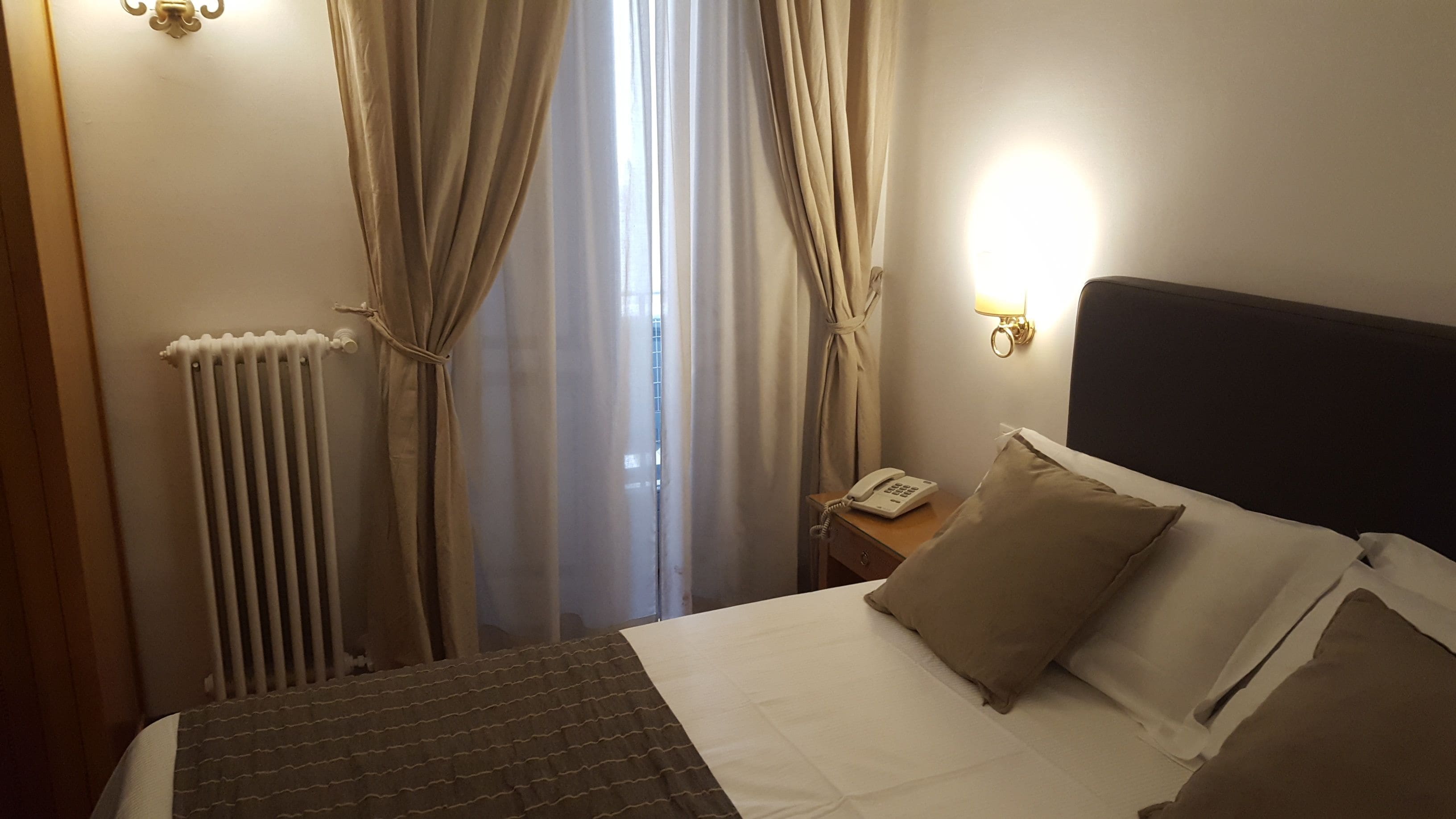Een hotelkamer met een bed, een nachtkastje, een lamp en een raam met gordijnen.