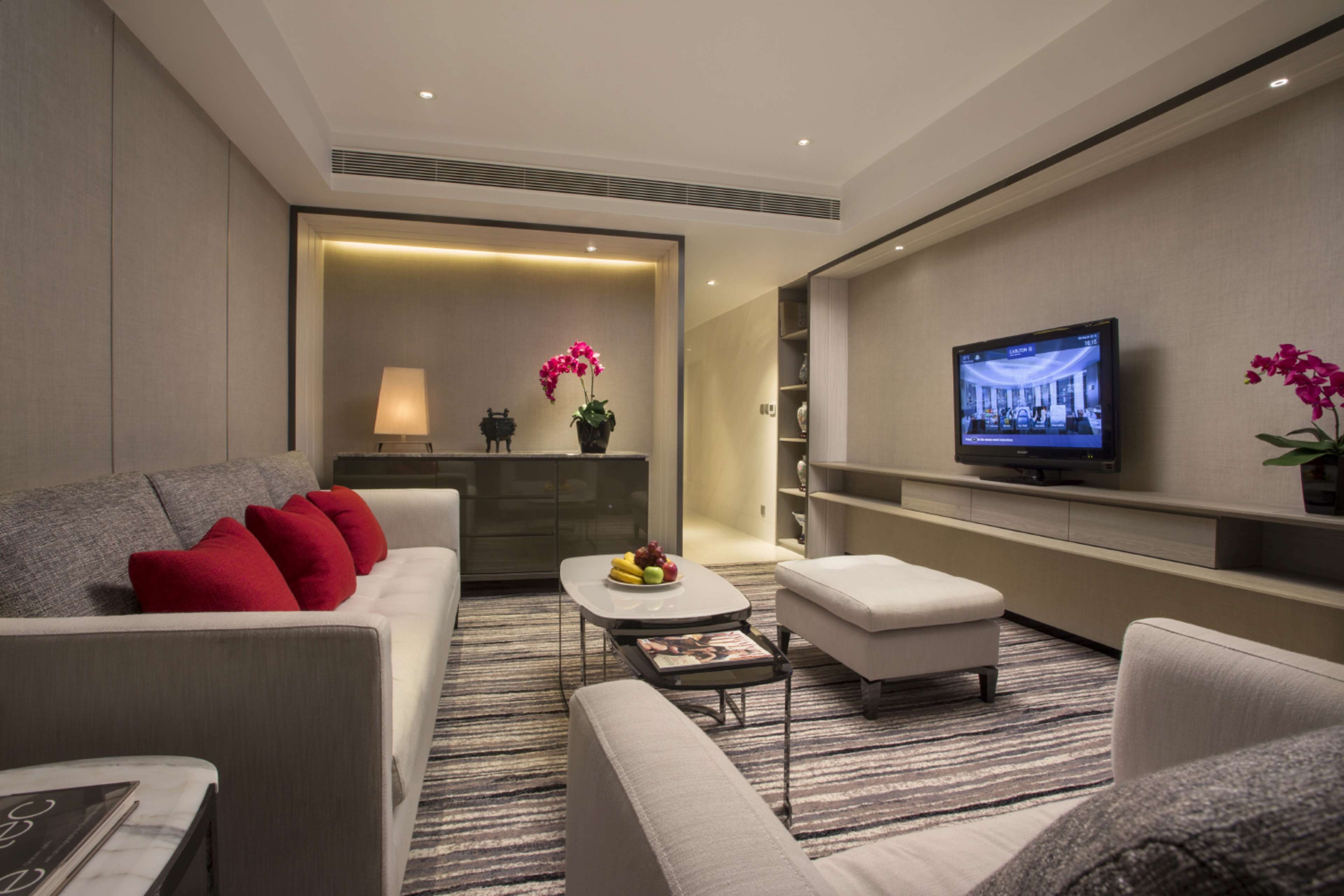A modern hotel room with a sofa, ottoman, flat-screen TV, and a small table with fruit.