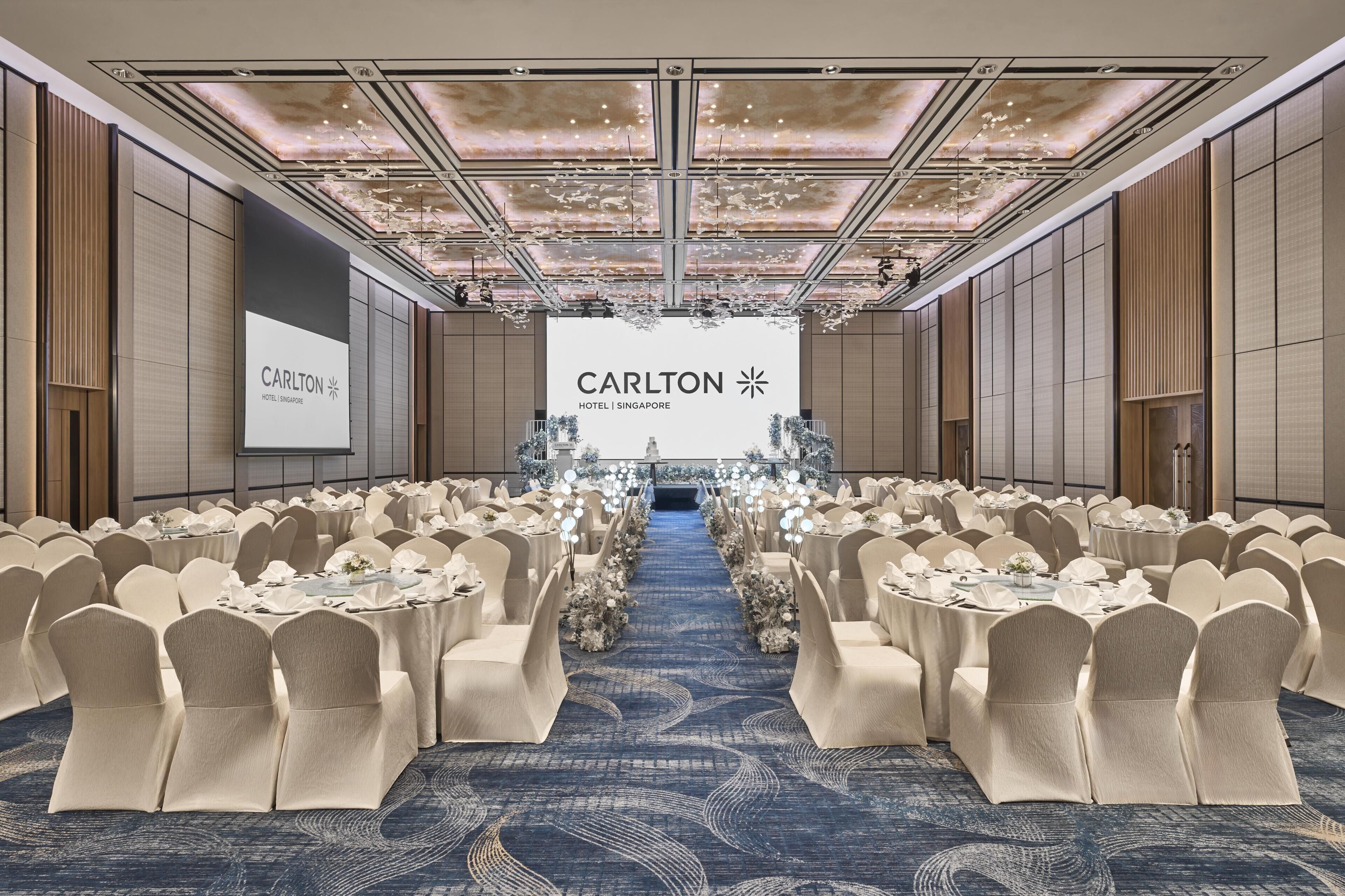 A conference room with round tables, beige chairs, and a large screen displaying 'Carlton Hotel Singapore'.