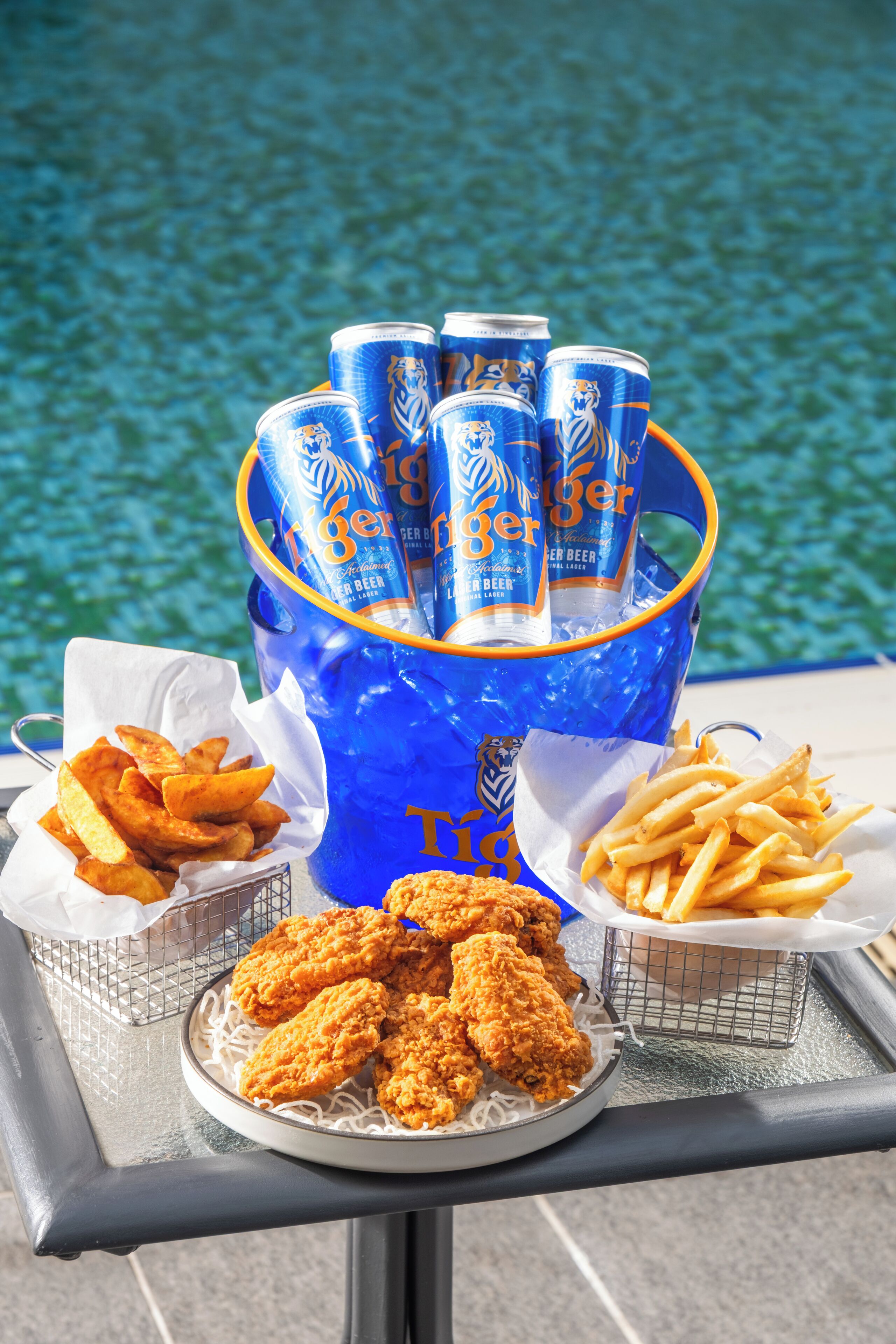 Una mesa con un cubo azul que contiene latas de cerveza Tiger, un plato de pollo frito y dos cestas de papas fritas.