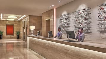 Uma área de recepção de hotel moderna, com um balcão de madeira, dois recepcionistas, uma parede de destaque vermelha e uma obra de arte contemporânea.