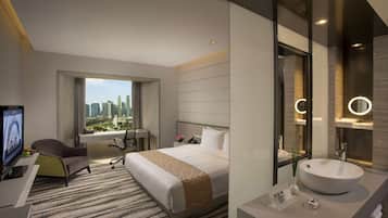 A hotel room with a large bed, a desk with a computer, a chair, a window with a city view, and a bathroom with a sink and mirror.