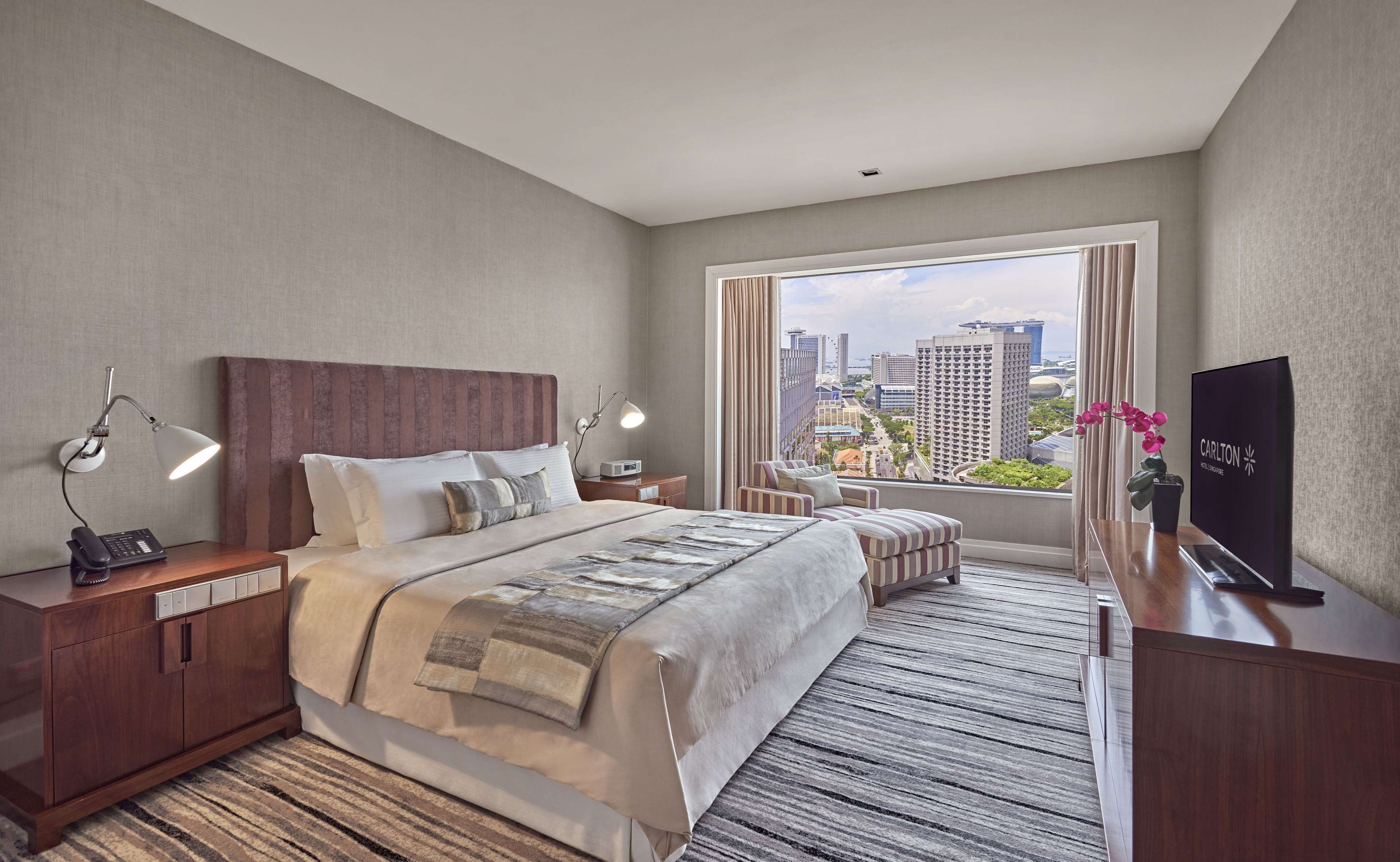 A hotel room with a large bed, bedside lamps, a desk with a phone, a TV, and a view of the cityscape.
