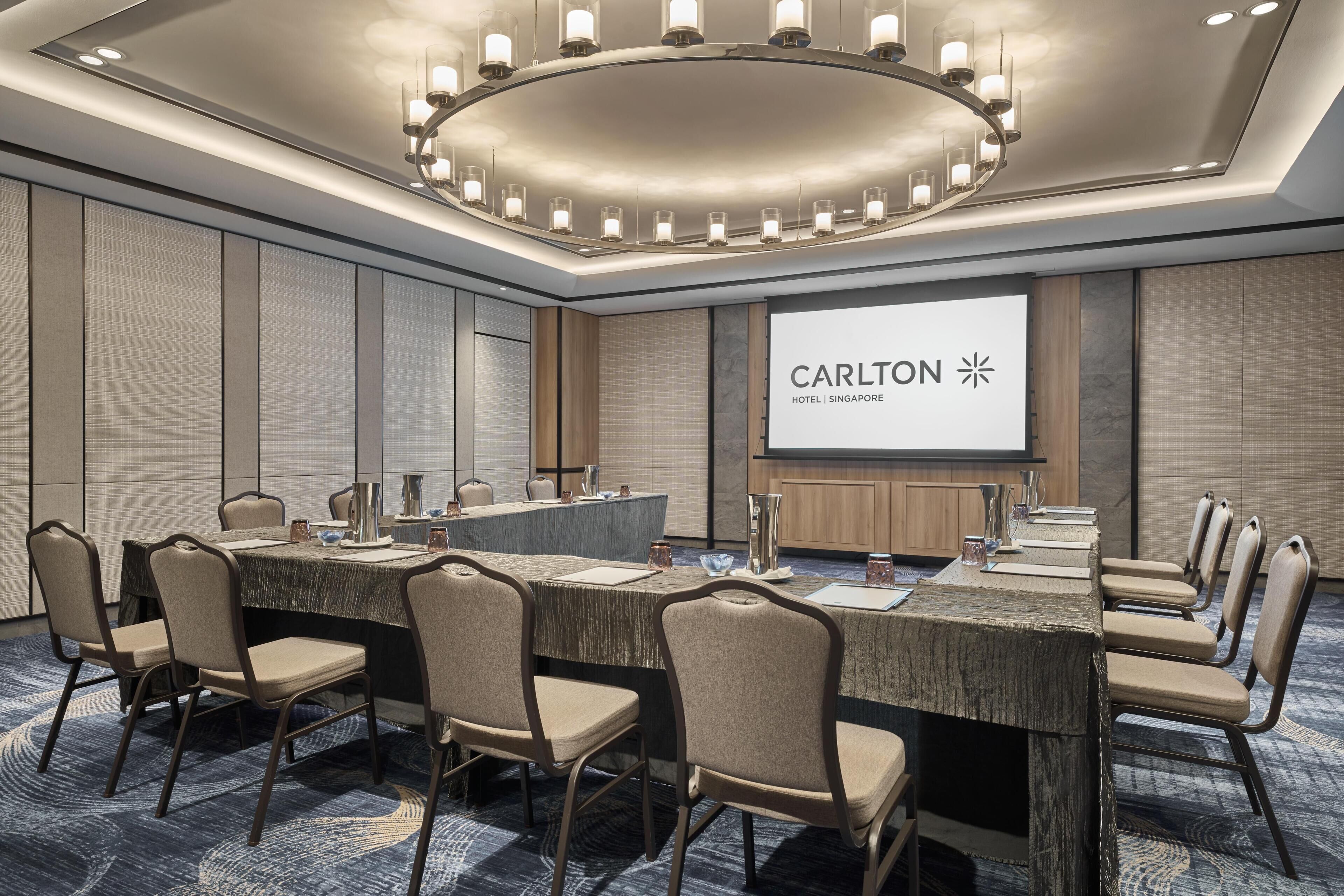 Una sala de conferencias con una mesa larga, sillas, una pantalla de proyector que muestra 'Carlton Hotel Singapore' y una lámpara de araña moderna.