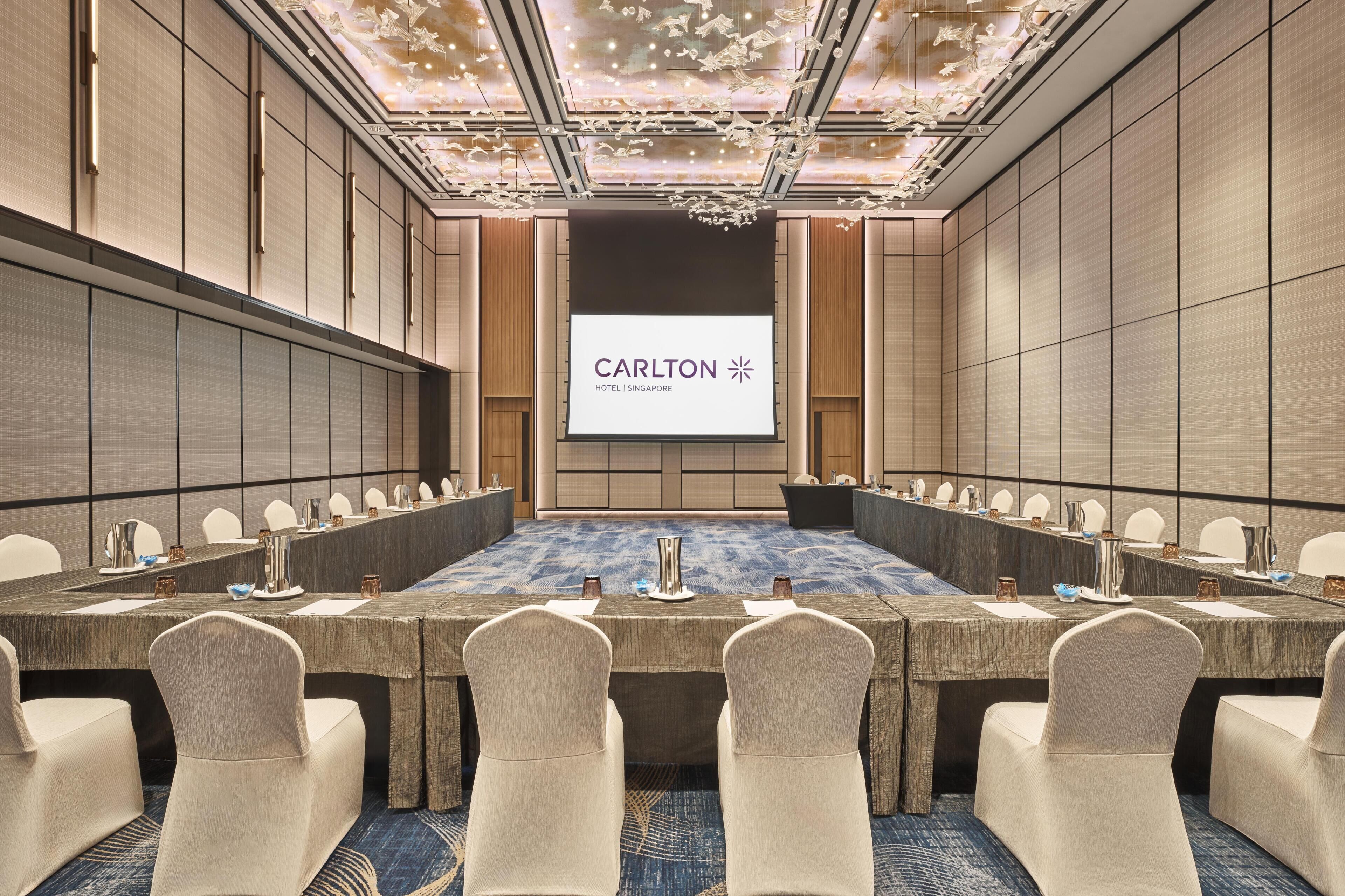 A conference room with a long table, chairs, and a screen displaying 'Carlton Hotel Singapore'.