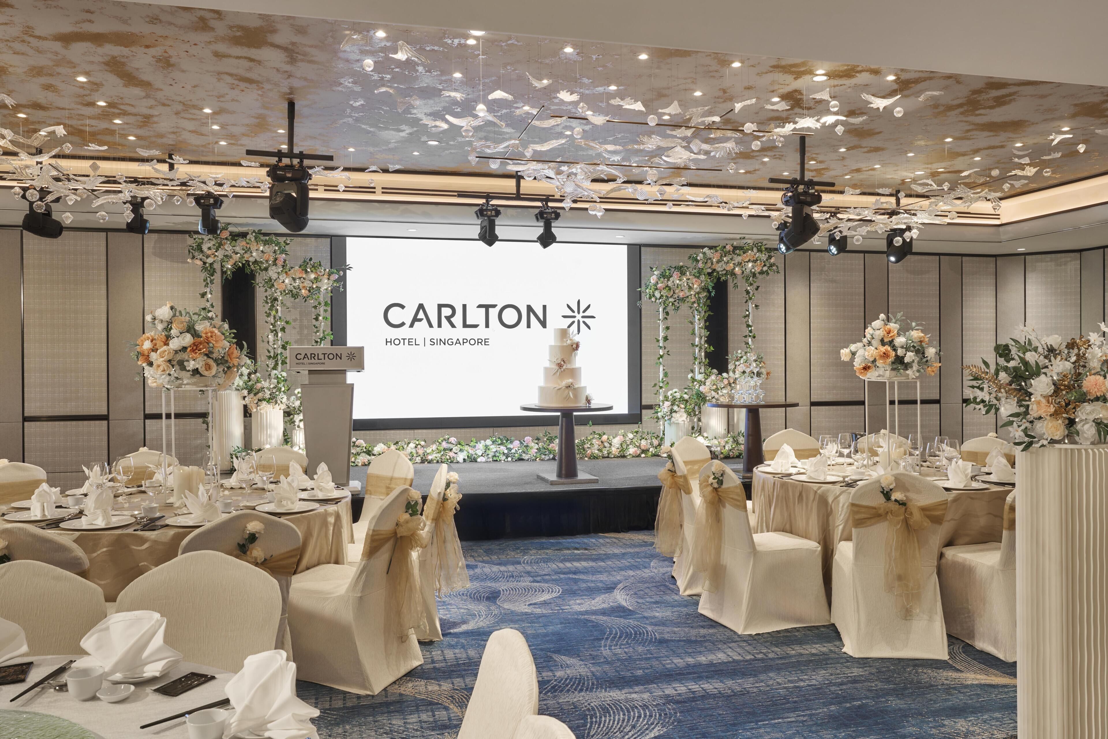 Una habitación de hotel con una pantalla grande mostrando el nombre del hotel, un escenario con un pastel y mesas preparadas para un evento formal.