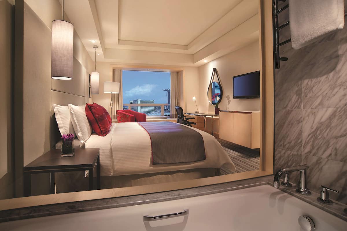 Une chambre d’hôtel avec un grand lit, un bureau, une chaise, une télévision et une salle de bain avec baignoire, offrant une vue sur la ville.
