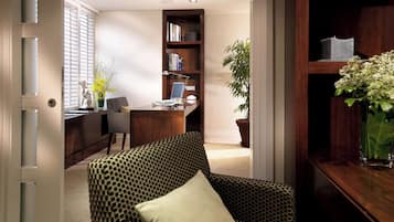 A room with a patterned armchair, a desk with a computer, a bookshelf, and a potted plant.
