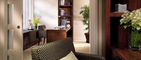 A room with a patterned armchair, a desk with a computer, a bookshelf, and a potted plant.