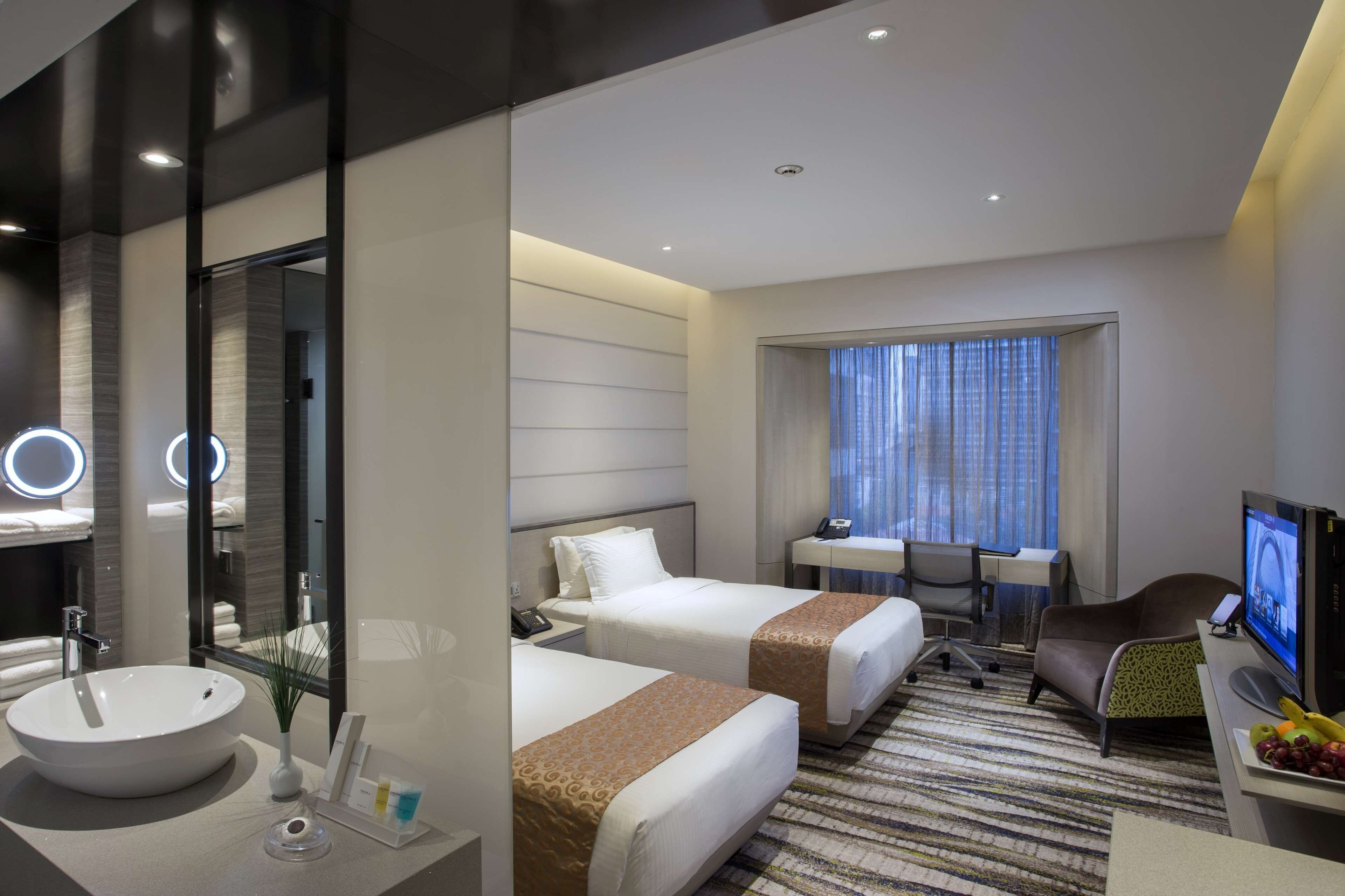 A hotel room with a large round sink, two beds, a desk with a chair, a television, and a window with curtains.