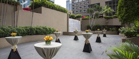 Um pátio com várias mesas redondas, algumas adornadas com flores amarelas, rodeado por vegetação exuberante e edifícios altos ao fundo.