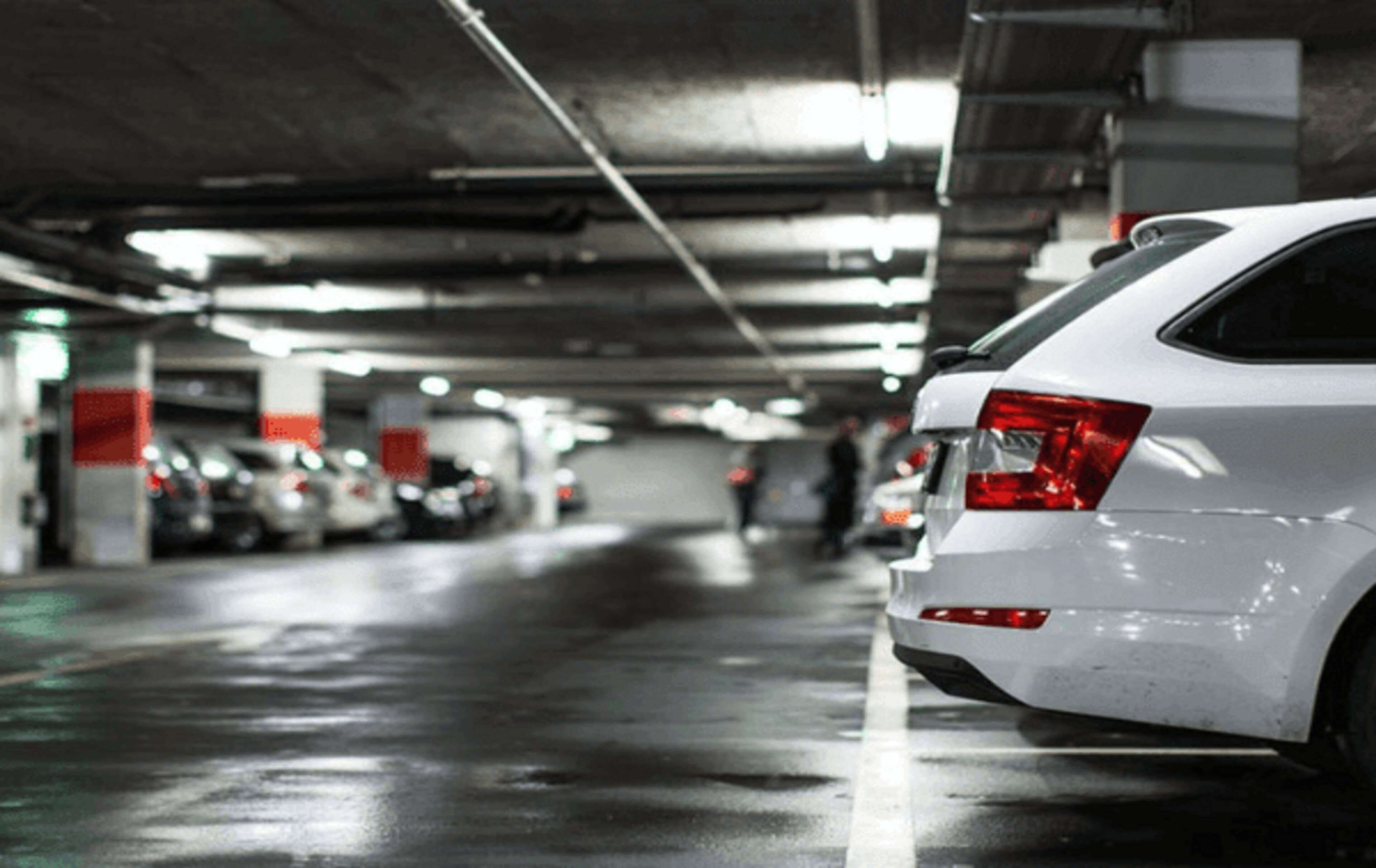 Ein weißes Auto steht in einer schlecht beleuchteten Tiefgarage mit Betonpfeilern und Deckenbeleuchtung.